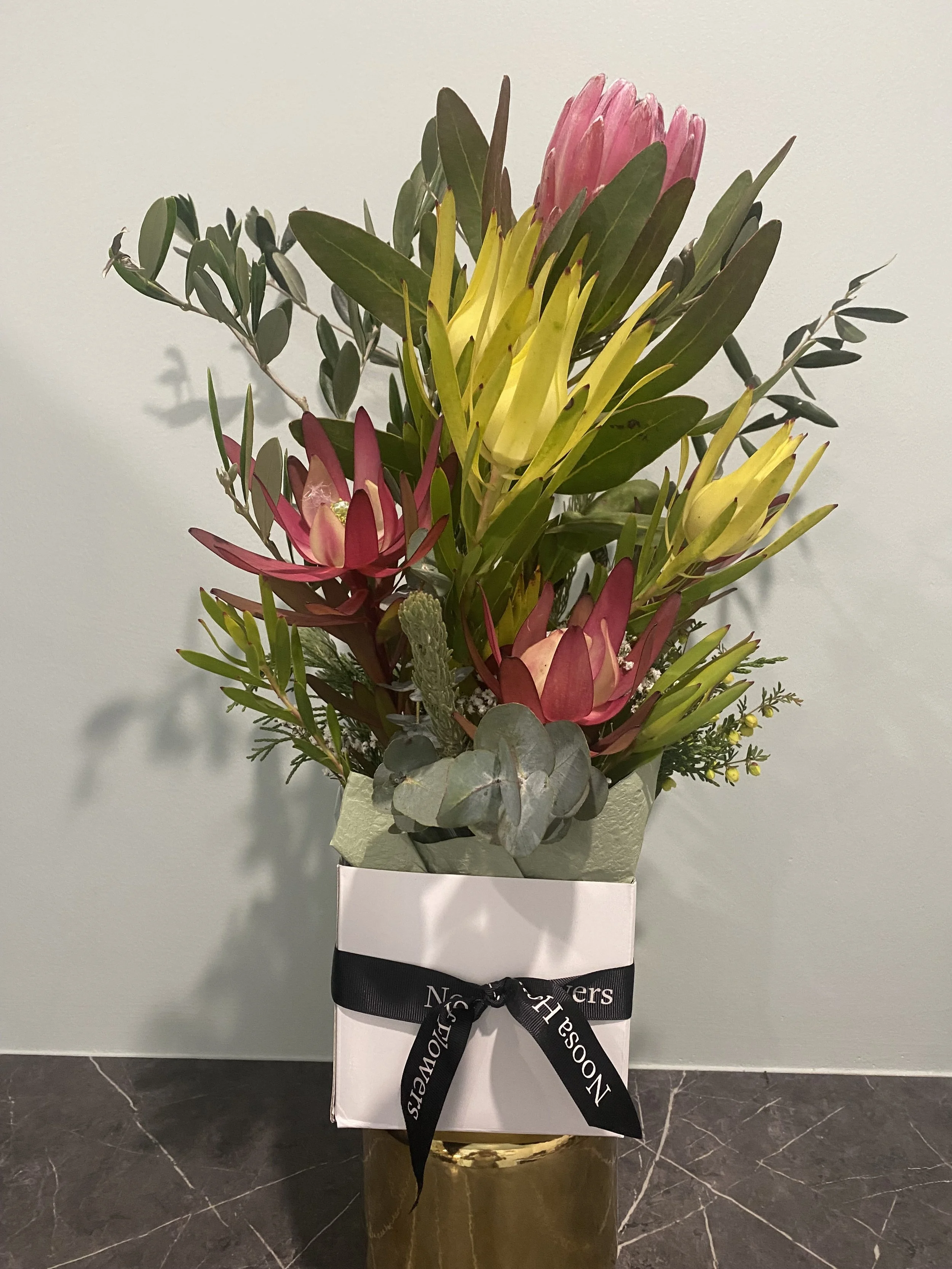 A floral arrangement in a white box with a black ribbon, featuring pink, yellow, and red flowers and greenery.