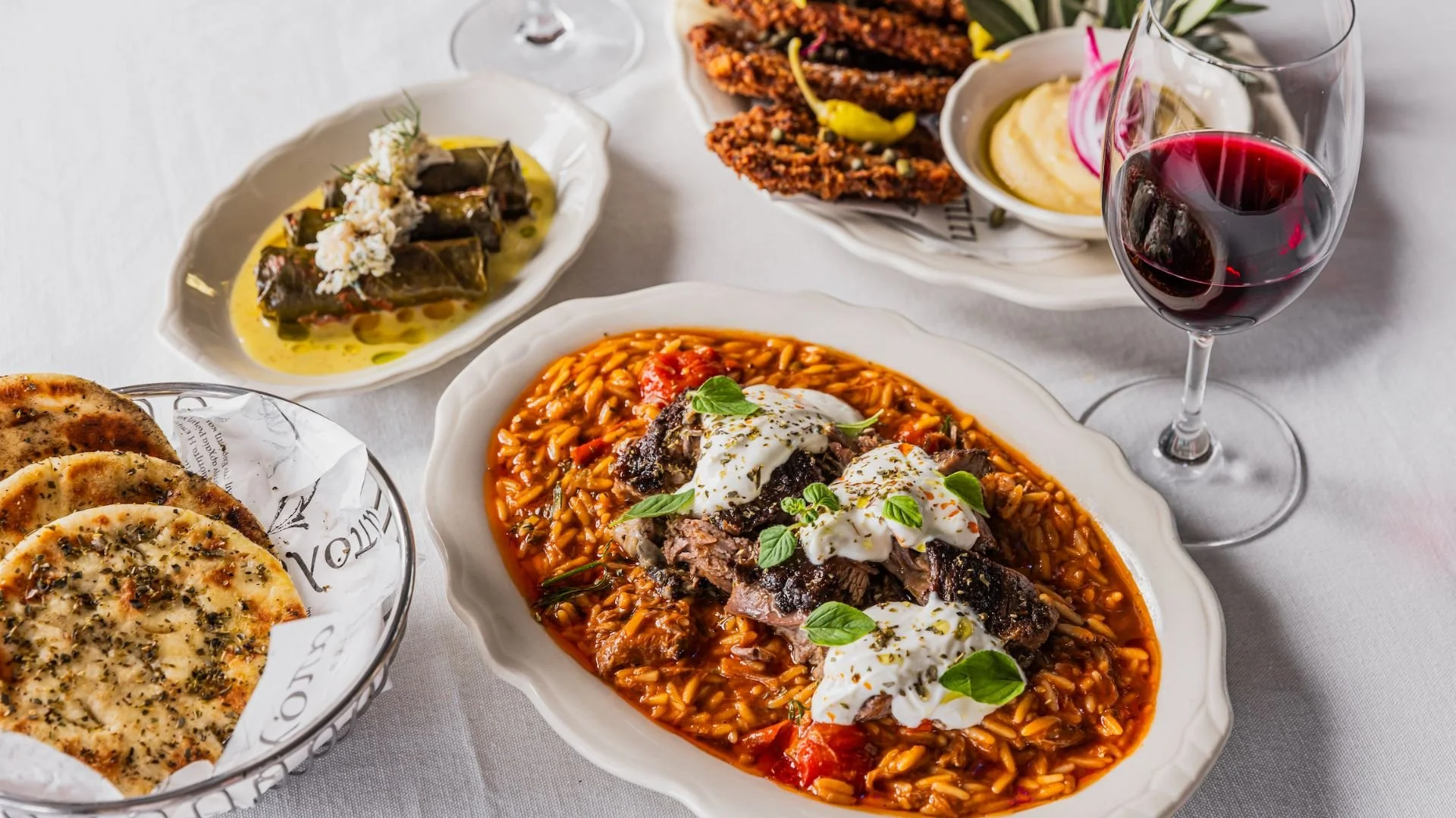 A table with various Greek dishes including a plate of braised beef or lamb with orzo pasta topped with dollops of Greek yogurt and fresh herbs, a bowl of OG mezze with spanakopita, tabbouleh, and other items, a basket of flatbread, and a glass of red wine.