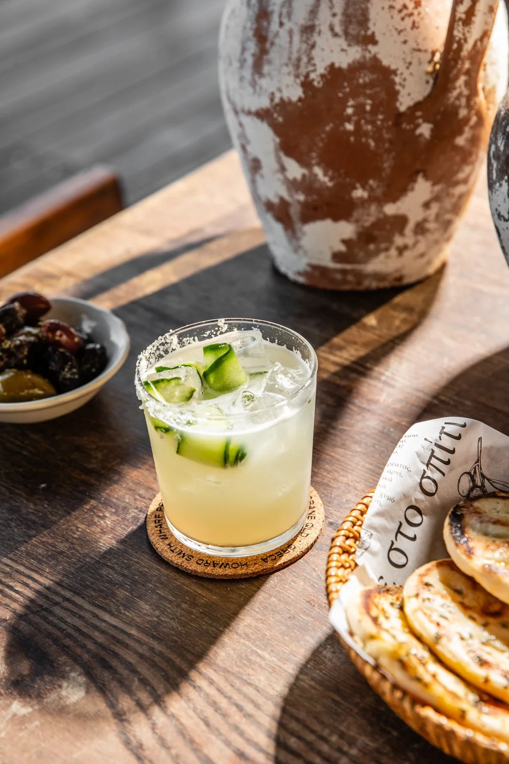 A glass of cucumber cocktail with ice on a wooden table, with a bowl of olives, a basket of grilled sandwiches, and large ceramic vases in the background.