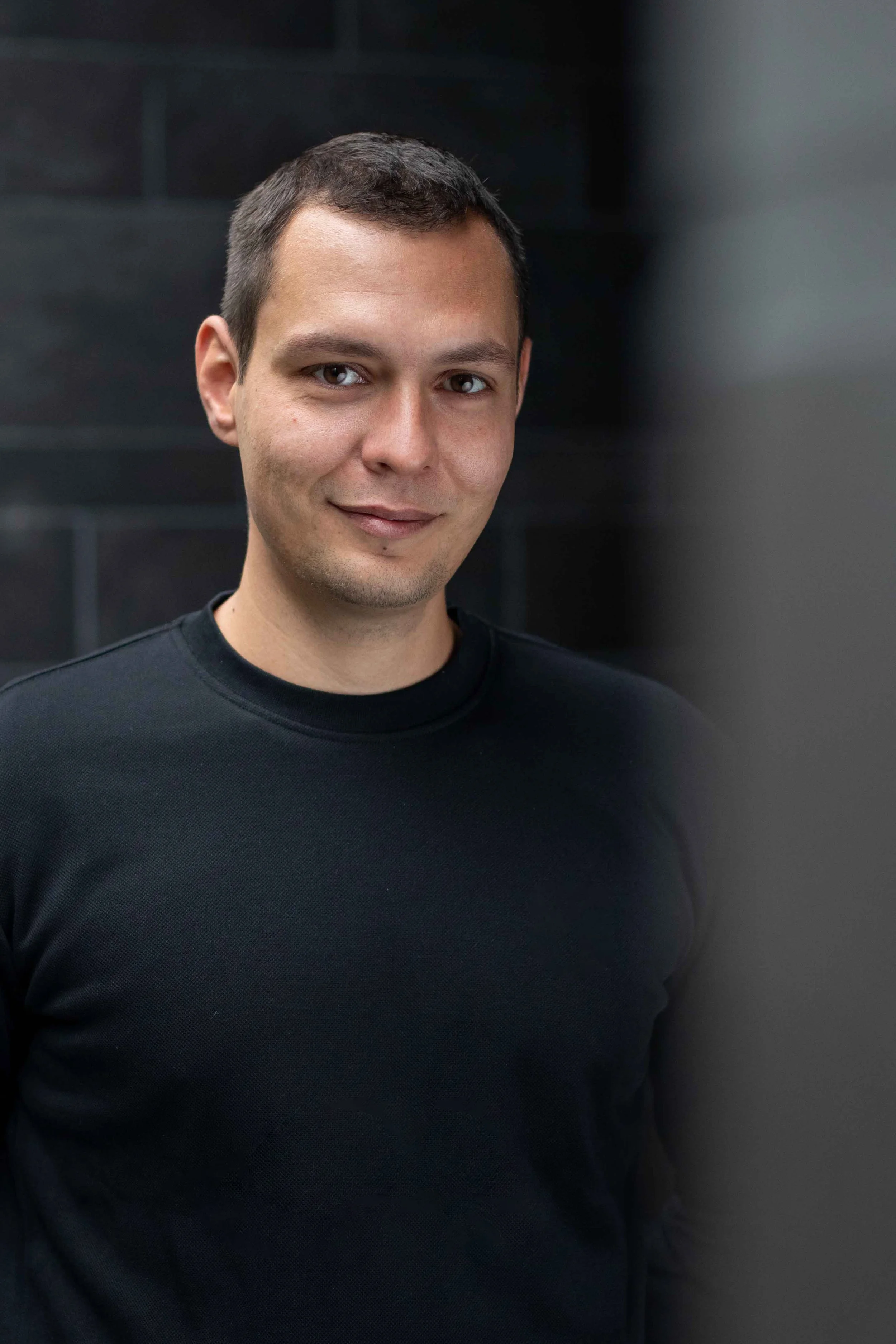 Junger Mann mit dunklem T-Shirt vor dunklem Hintergrund, leicht lächelnd, in Nahaufnahme.