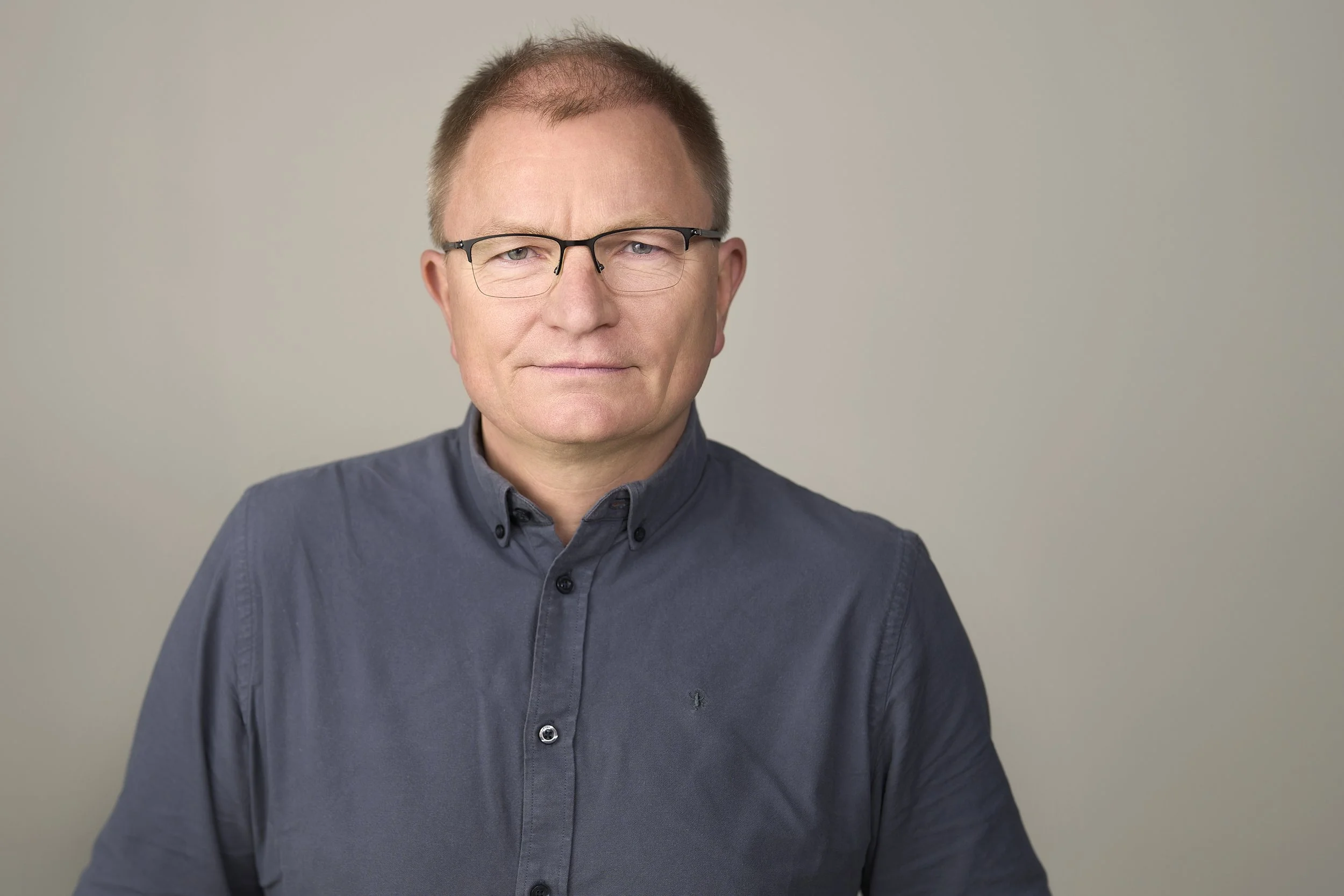Portrett av en mann med briller i en mørk grå skjorte mot en lys grå bakgrunn.