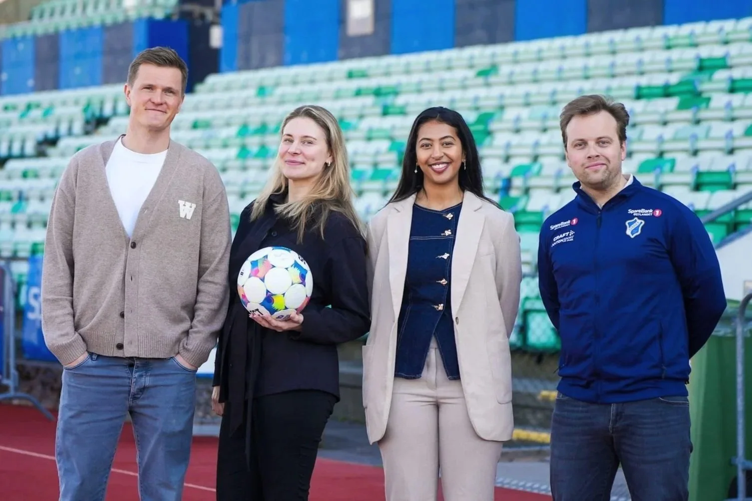 Fire menn og to kvinner står på en fotballarena, en av kvinnene holder en fargerik fotball. De smiler og poserer for fotografen.