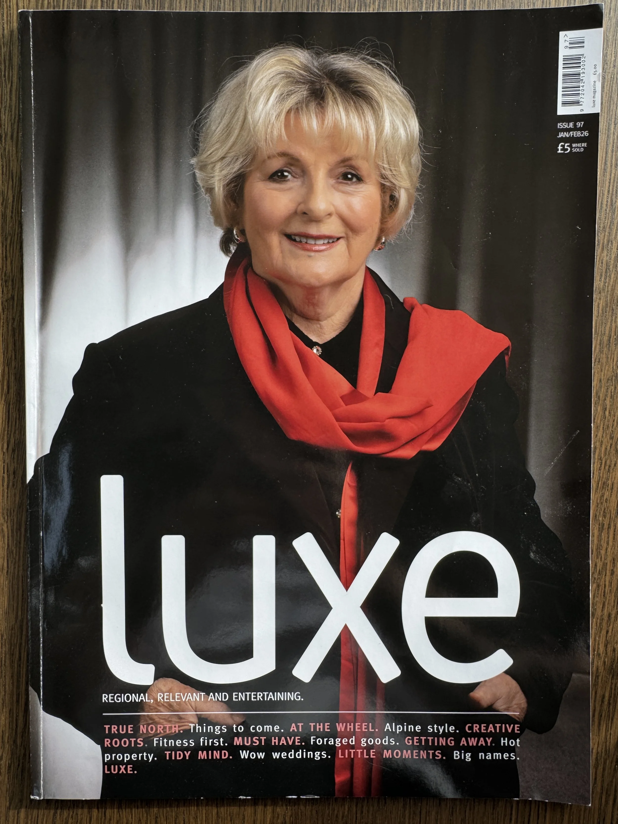 A woman with short blonde hair smiling, dressed in a black outfit with a red scarf, on the cover of Luxe magazine.