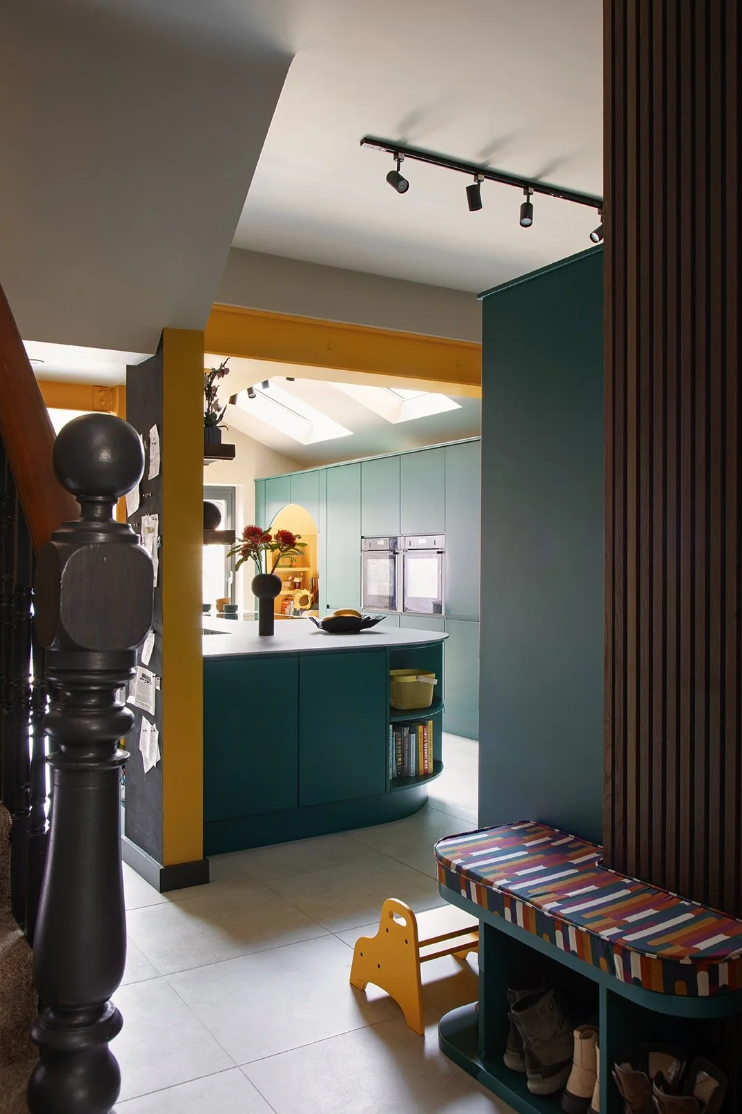 View from the hallway into the open-plan kitchen of a North Shields family home by Carole Crowe Interior Design, framed by a painted black Victorian newel post and dark timber slatted wall panel, showing the curved teal kitchen island with open cookb