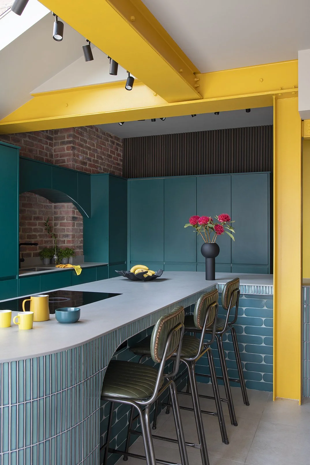 Wide view of the open-plan kitchen in a North Shields family home by Carole Crowe Interior Design, showing the curved teal glazed tile island with a grey stone worktop, olive green leather studded bar stools, a yellow painted structural steel portal 