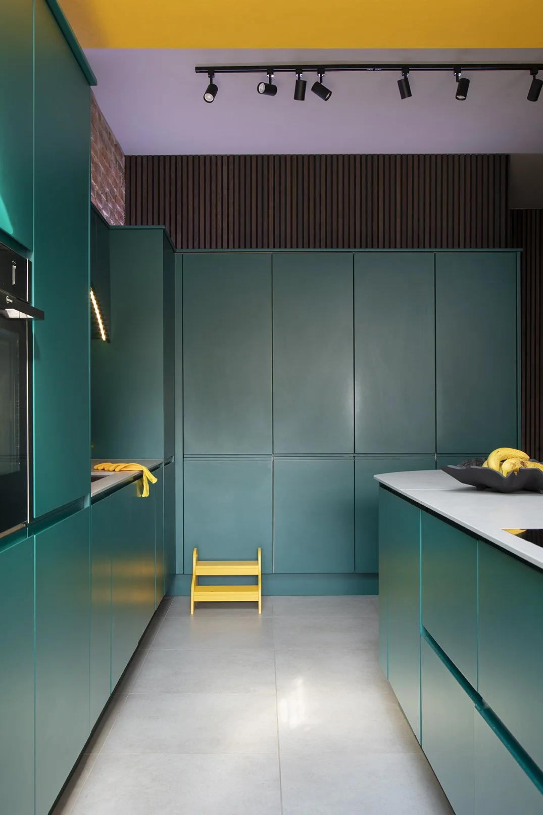 View along the kitchen run in a North Shields family home designed by Carole Crowe Interior Design, showing floor-to-ceiling teal handleless cabinetry against a lilac painted ceiling with a yellow painted structural beam above, dark timber acoustic s