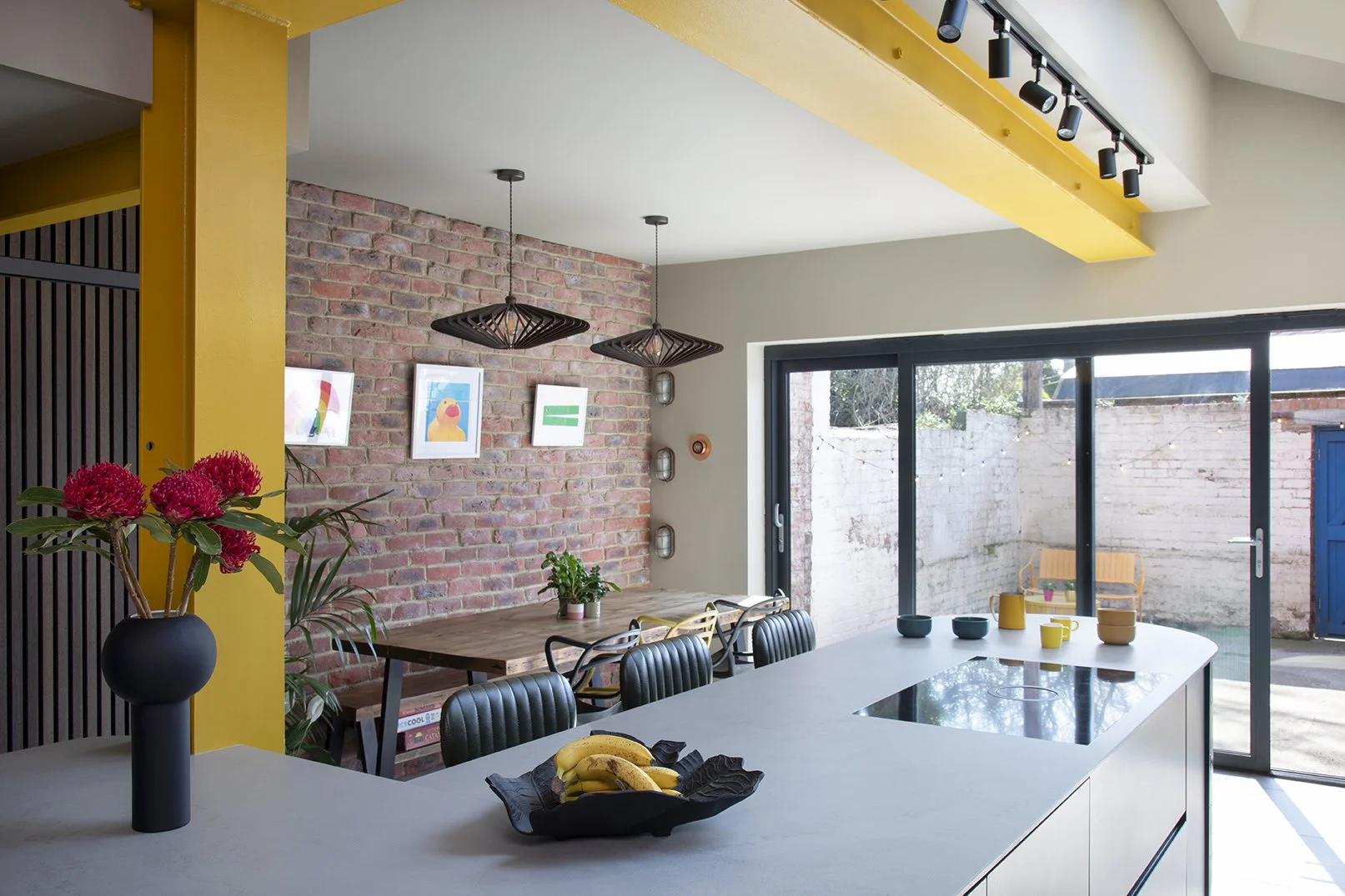Wide view of the open-plan kitchen and dining room in a North Shields family home designed by Carole Crowe Interior Design, showing the curved teal tiled kitchen island with grey stone worktop, a matte black vase with waratah flowers, a solid wood di