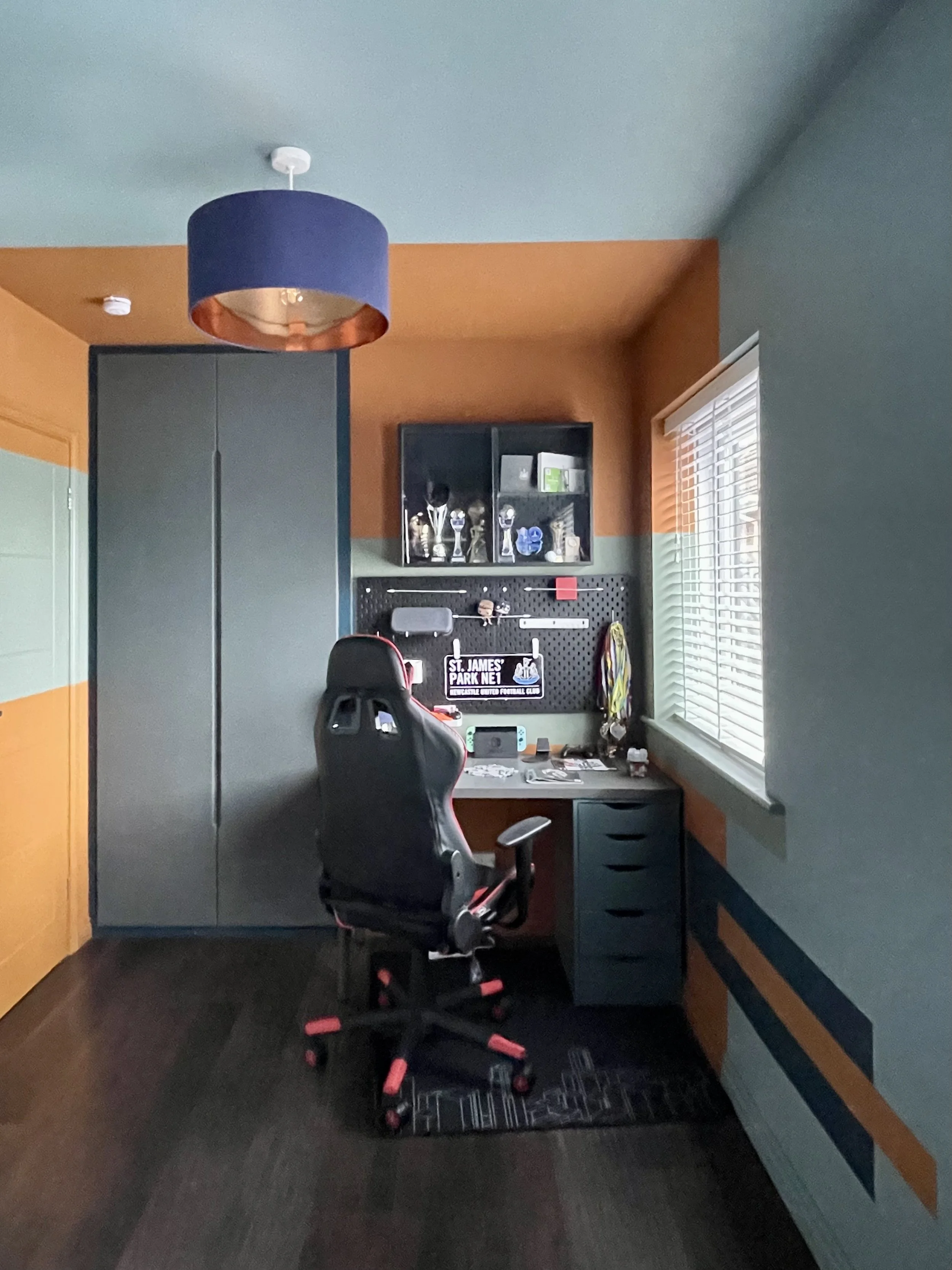 Full boys' bedroom showing gaming desk, built-in wardrobe, colour-blocked orange and teal walls and navy copper pendant