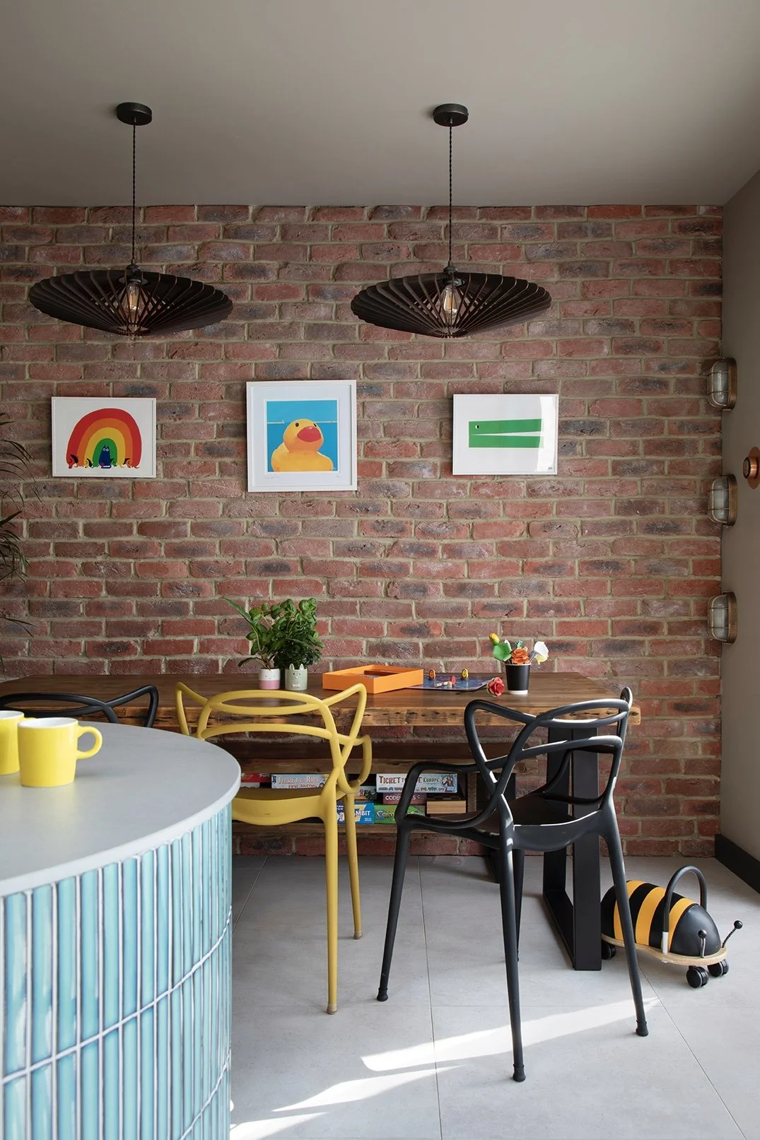 Family dining area in a North Shields home designed by Carole Crowe Interior Design, featuring a solid wood dining table with yellow and black Masters-style chairs, three framed children's artworks hung on an exposed red brick wall, two oversized bla