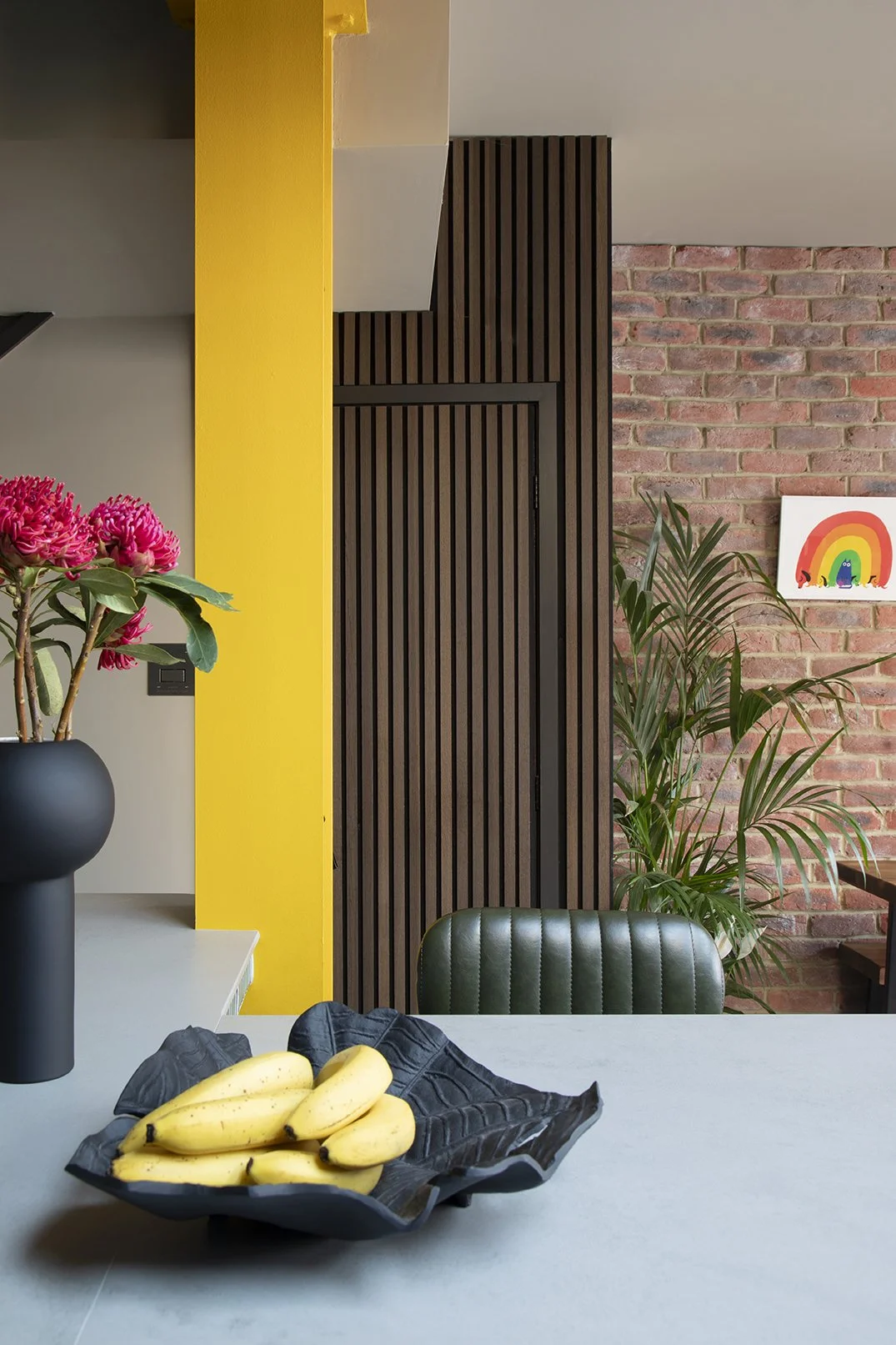 Evening lighting view of a bold family kitchen by Carole Crowe Interior Design in North Shields, showing teal handleless cabinetry, a curved teal tiled kitchen island with grey stone worktop, yellow painted structural steel beam, warm ambient ceiling