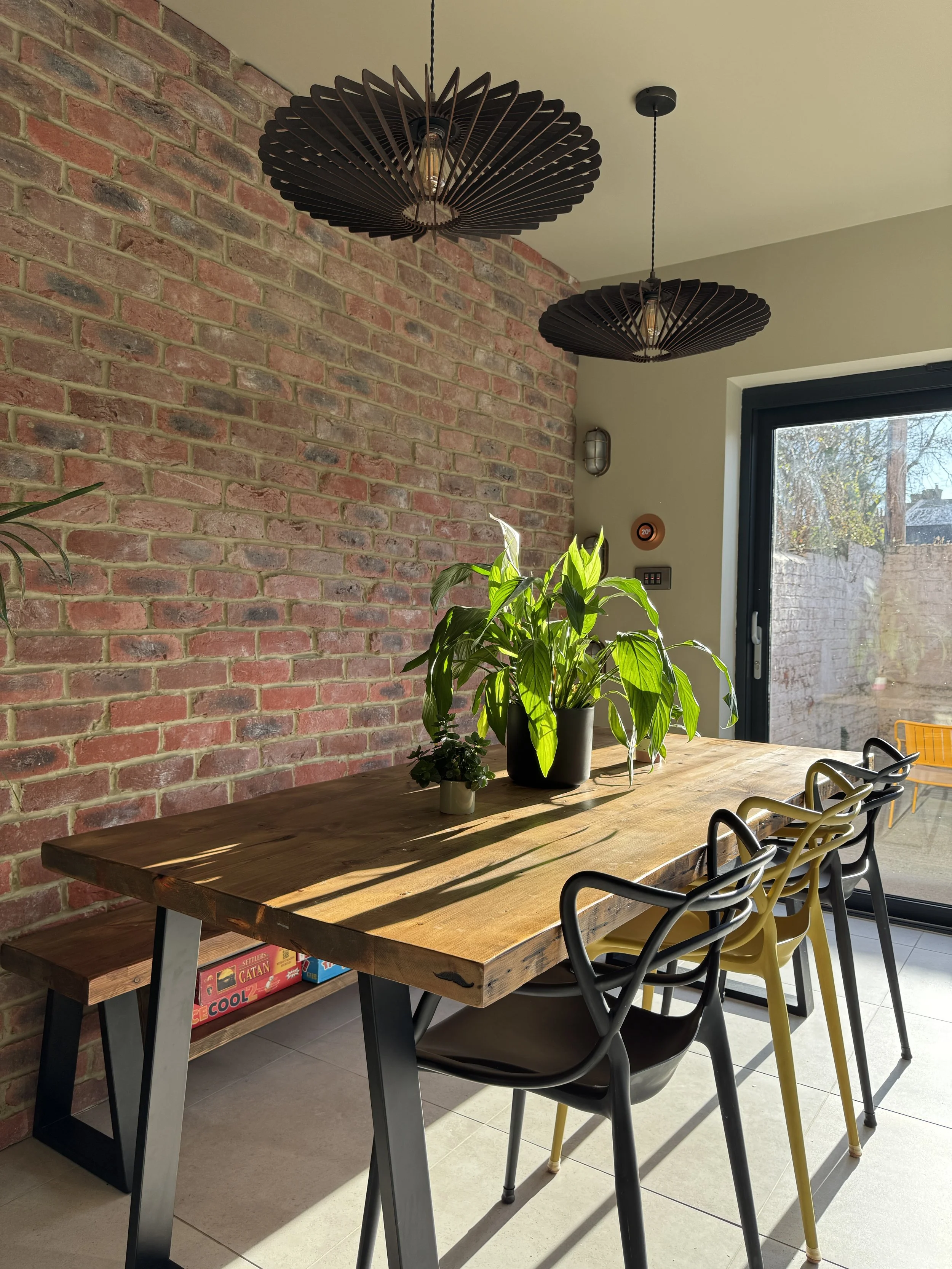 Sunlit dining area in a North Shields family home designed by Carole Crowe Interior Design, featuring a reclaimed solid wood dining table with steel A-frame legs, black and yellow Masters-style chairs, two large black sculptural pendant lights, an ex