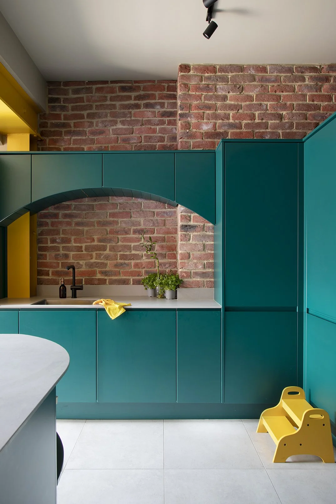 Kitchen sink area in a North Shields family home by Carole Crowe Interior Design, featuring teal handleless base and larder cabinetry with a grey stone worktop, a black matt tap and undermount sink, an arched teal-framed opening revealing an exposed 