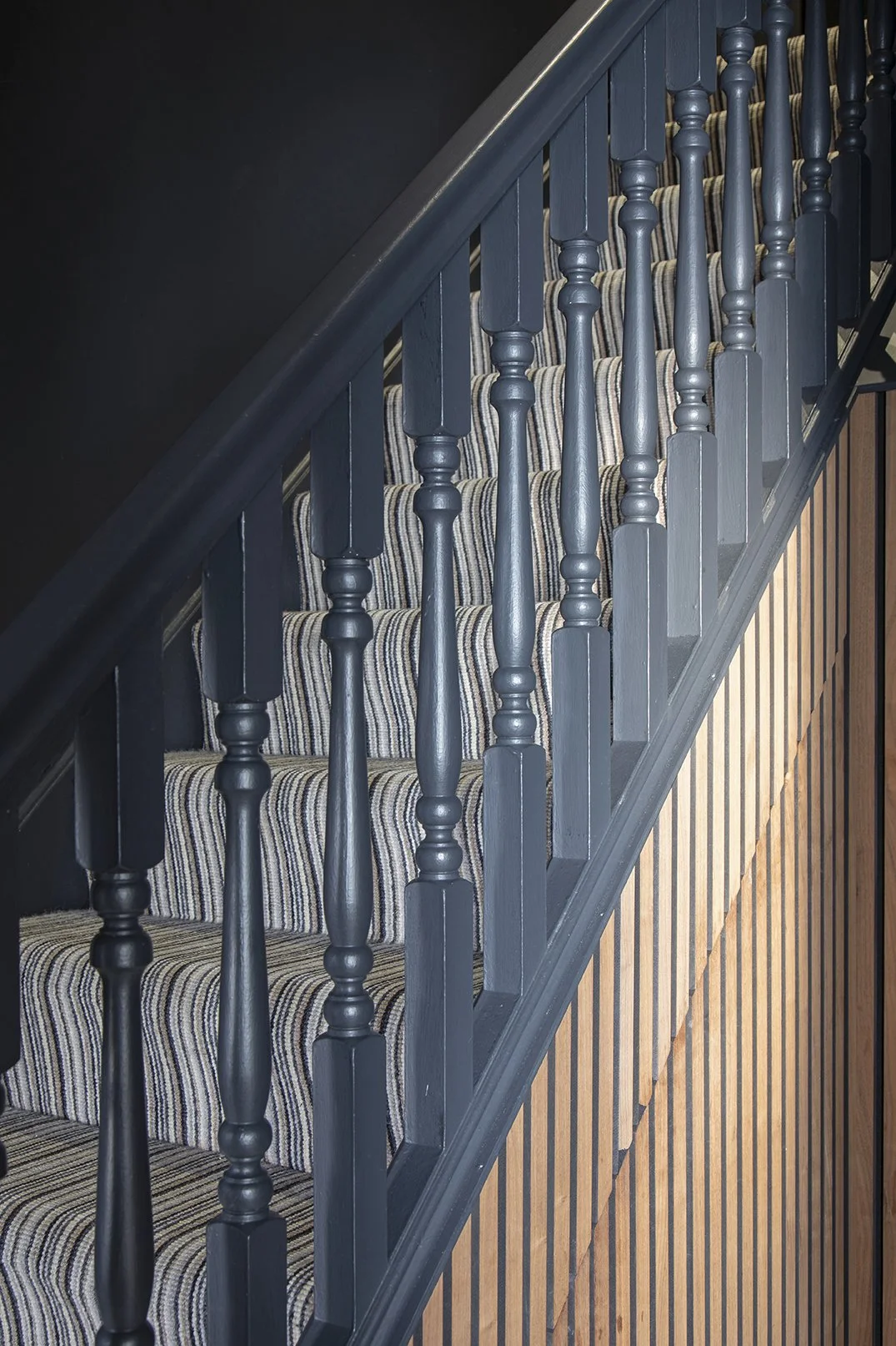 Close-up of painted Black blue staircase spindles with striped carpet runner and warm oak wood slat panel, Tynemouth