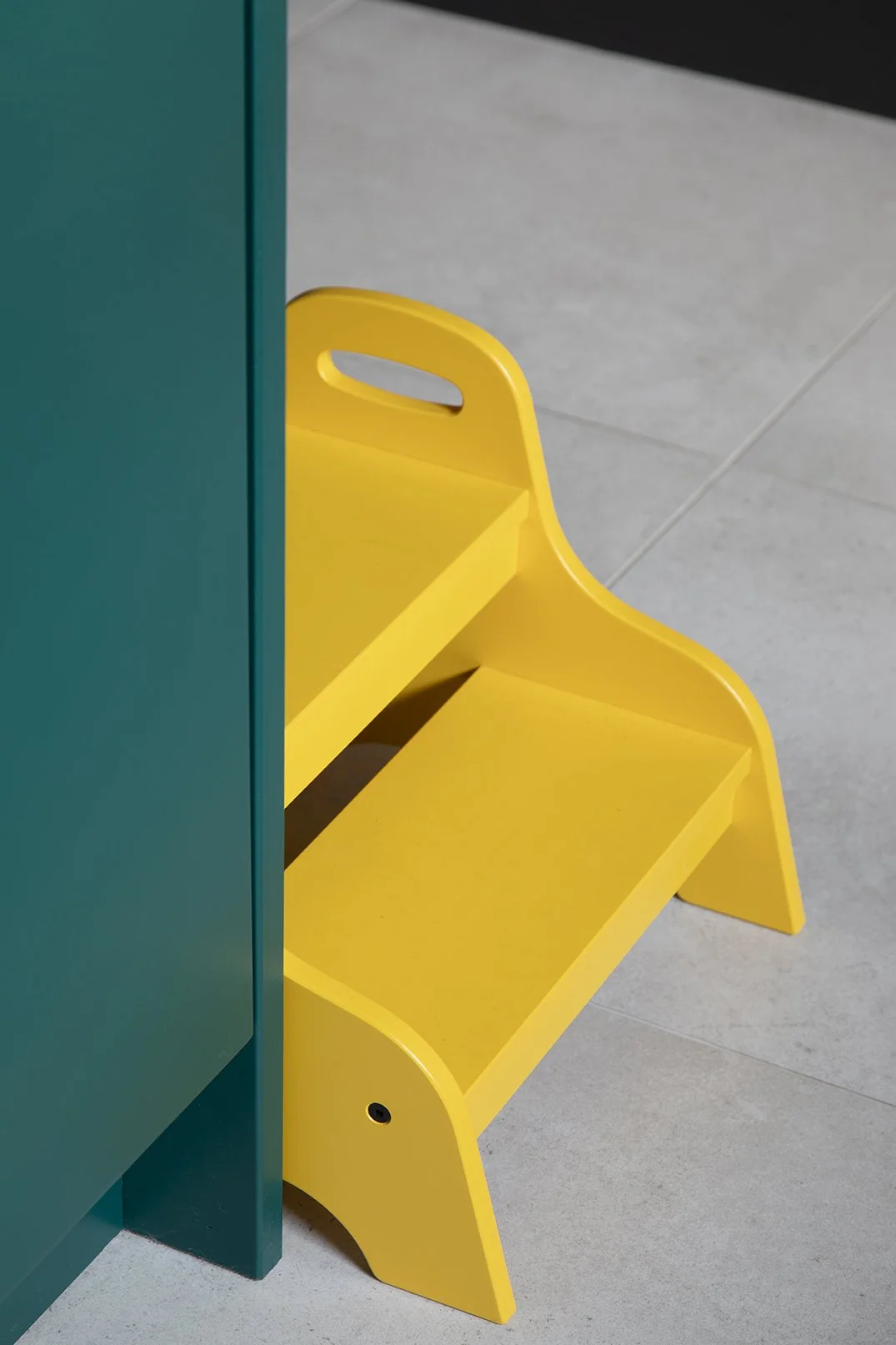 Close-up detail of a bright yellow children's step stool positioned beside a teal kitchen cabinet on large format porcelain tile flooring in a North Shields family home designed by Carole Crowe Interior Design, illustrating the playful, considered ap