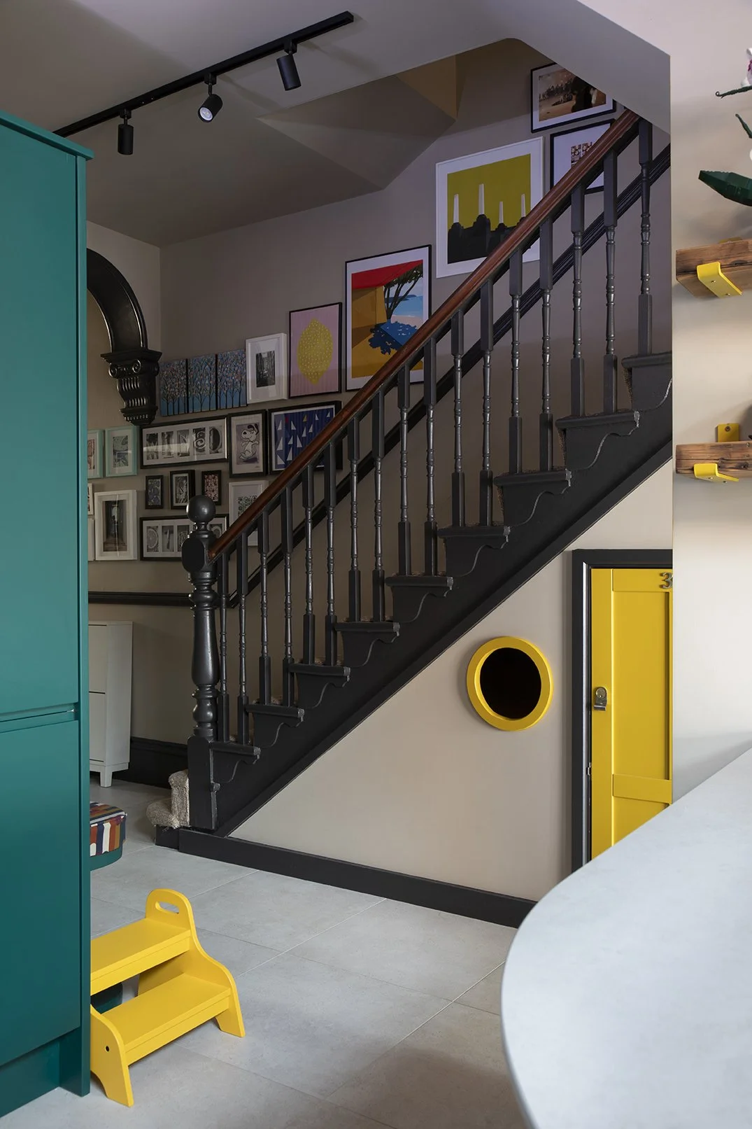 Staircase hall in a North Shields family home by Carole Crowe Interior Design, showing a painted black Victorian staircase, a curated gallery wall of framed prints and original artworks climbing the stairs, a yellow under-stairs children's playroom d