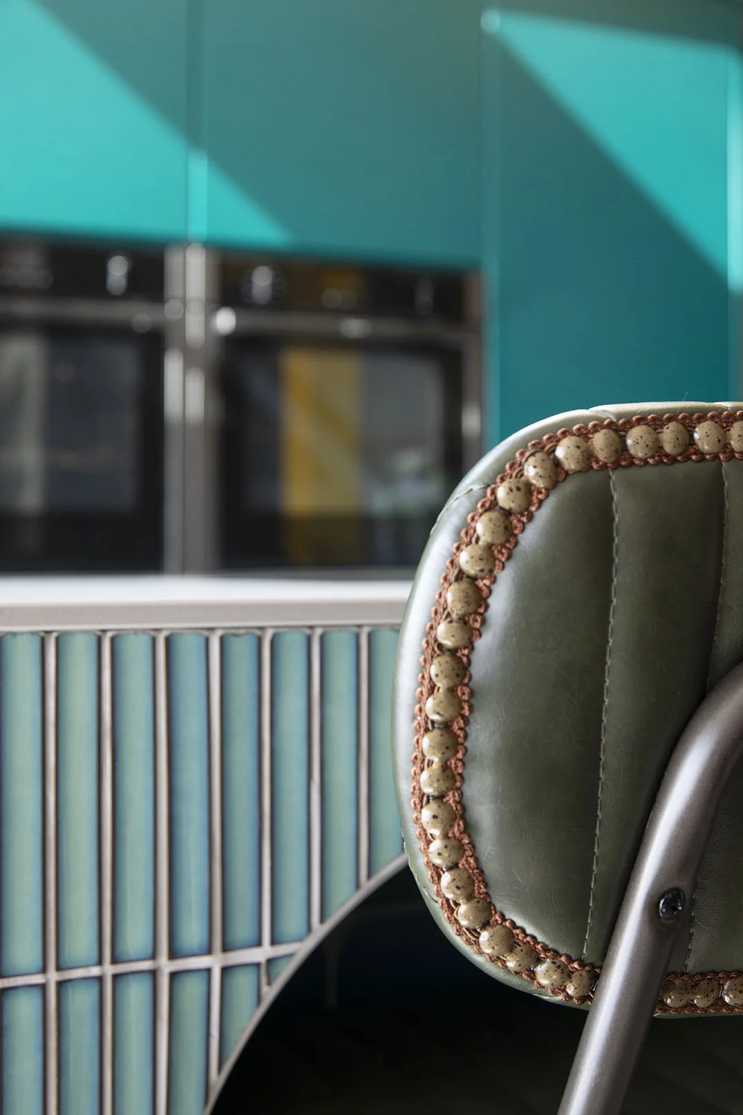 Close-up detail of an olive green leather bar stool with decorative studded trim set against the curved teal glazed finger tile kitchen island in a North Shields family home designed by Carole Crowe Interior Design, with teal handleless cabinetry and