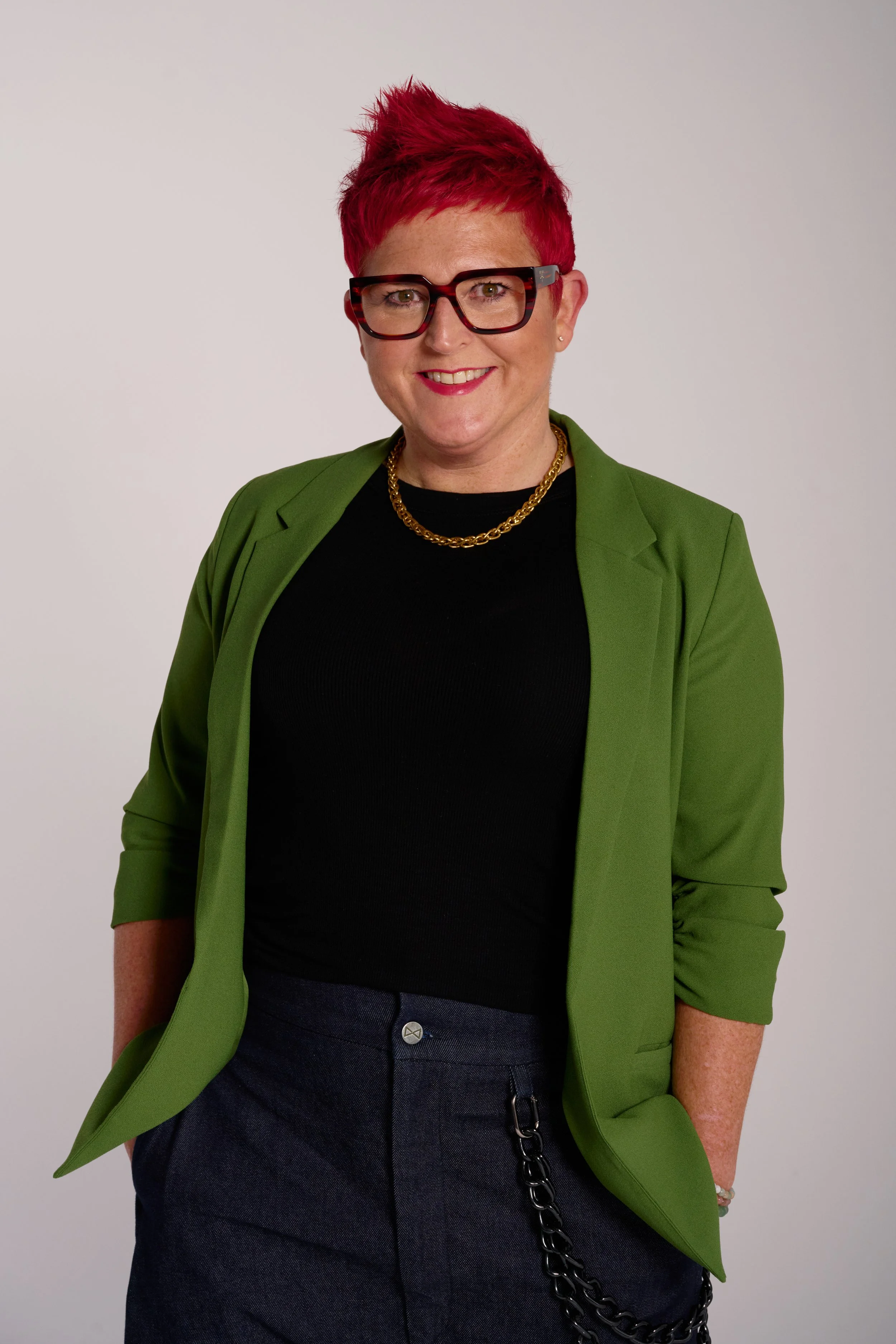 A woman with short red hair, wearing large tortoiseshell glasses, a black top, a green blazer, dark pants, a gold chain necklace, and a chain accessory hanging from her pocket, standing against a plain light gray background and smiling at the camera.