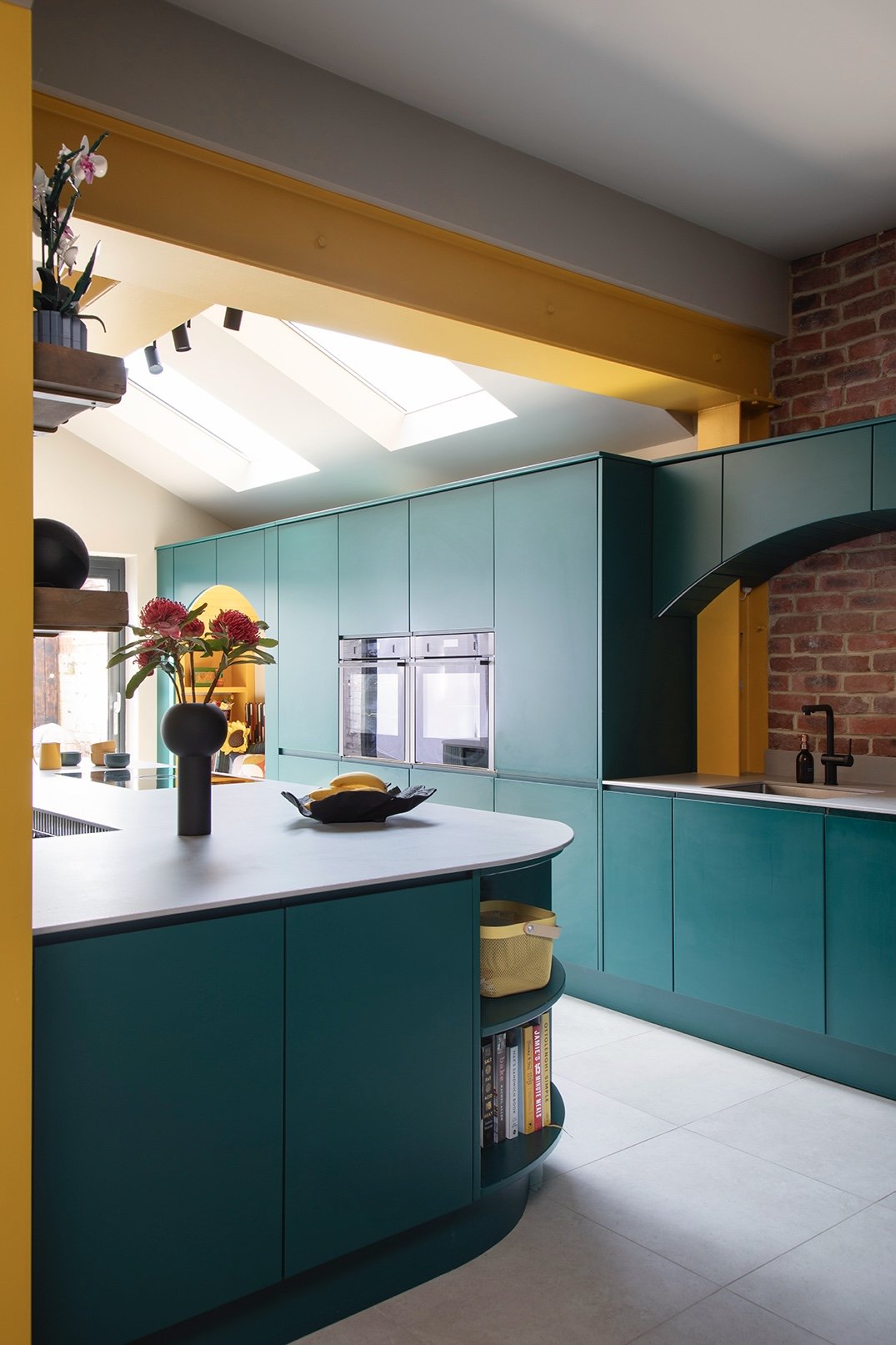 Bold family kitchen designed by Carole Crowe Interior Design in North Shields, showing a curved teal handleless kitchen island with open shelving, a run of teal floor-to-ceiling cabinetry with integrated double ovens, an arched teal-framed recess rev