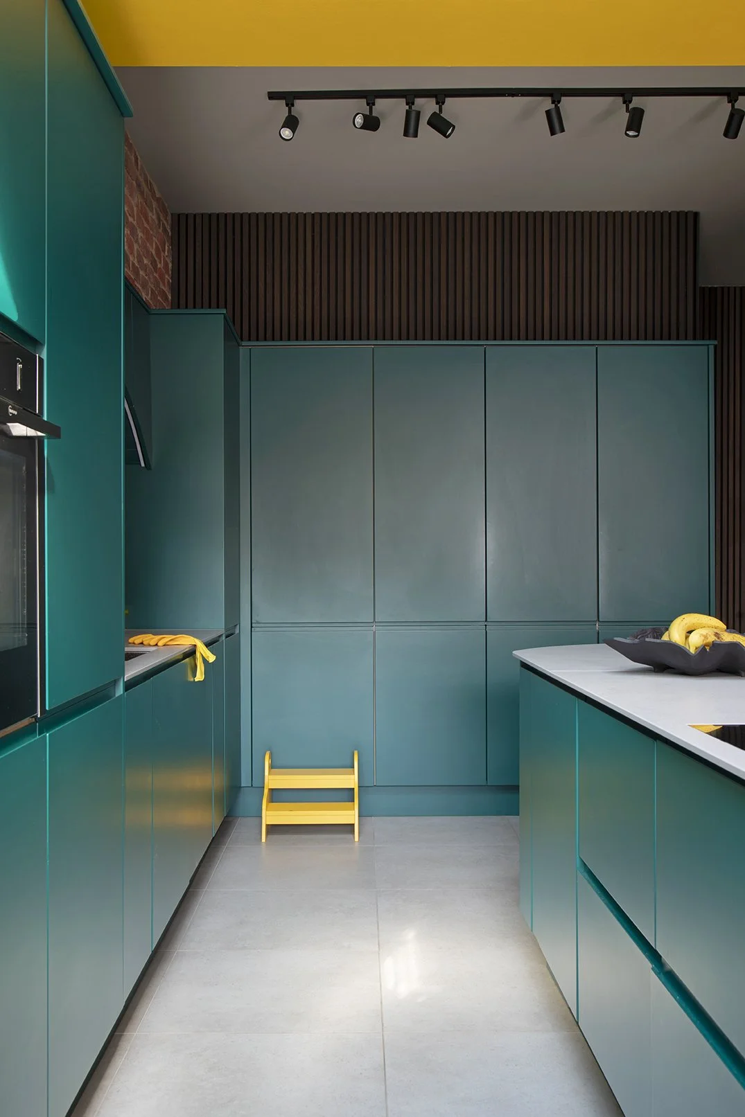 View along the kitchen run in a North Shields family home designed by Carole Crowe Interior Design, showing floor-to-ceiling teal handleless cabinetry with fully concealed appliance cupboards closed, a yellow painted ceiling, dark timber acoustic sla