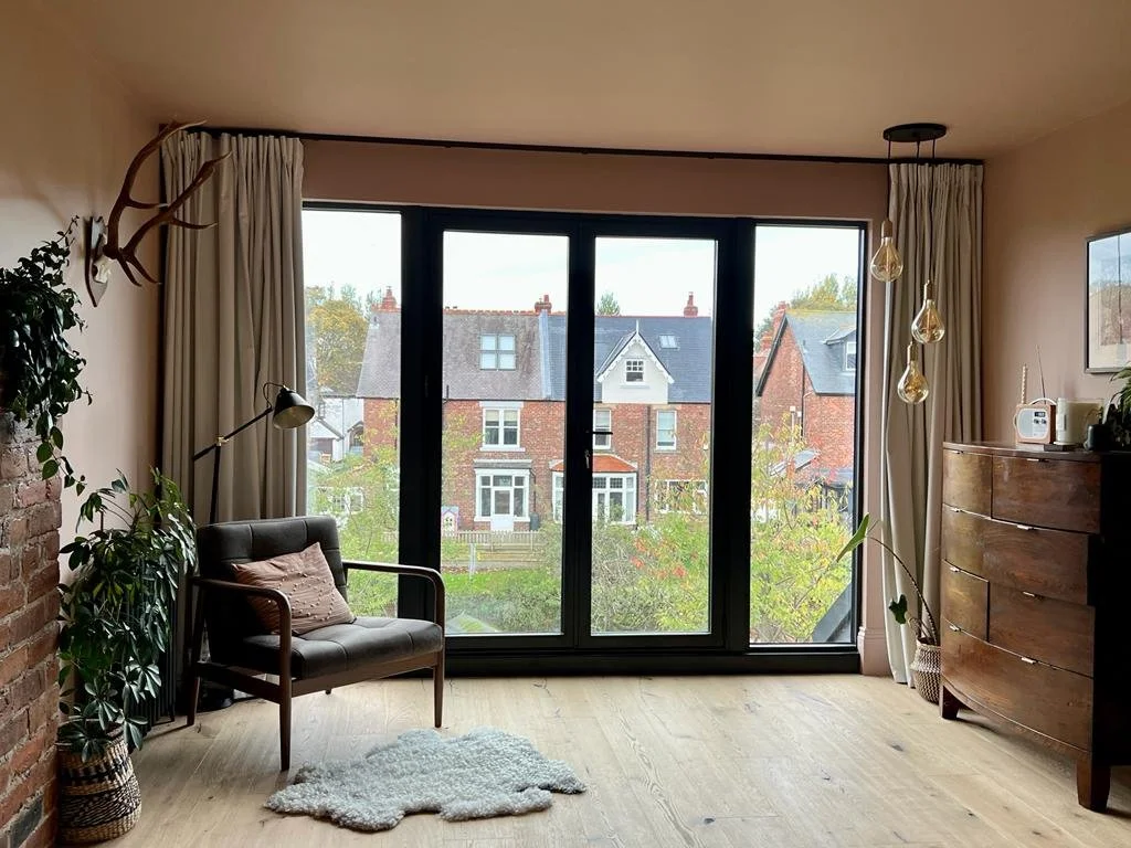 Completed loft bedroom in the Queens Road project by Carole Crowe Interior Design, Whitley Bay, featuring blush pink painted walls, wide-plank light oak flooring, large black-framed picture windows with linen curtains overlooking a residential street