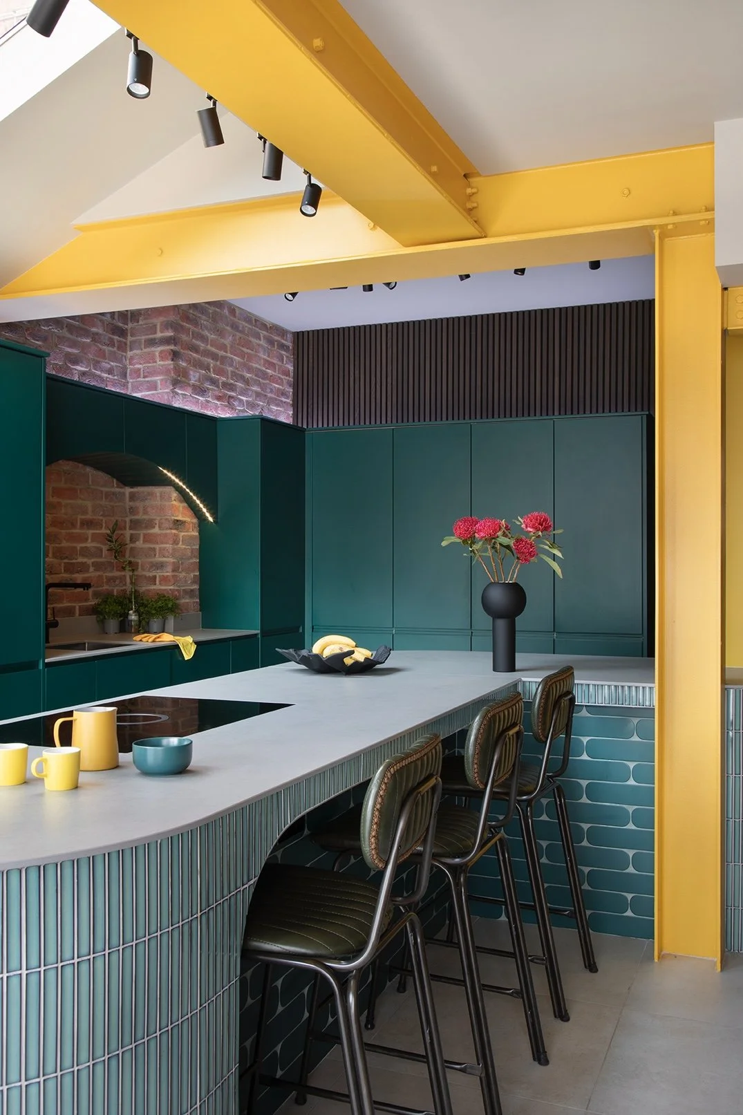 Evening lighting view of the open-plan kitchen in a North Shields family home designed by Carole Crowe Interior Design, featuring the curved teal tiled island with grey stone worktop, olive green leather studded bar stools, a bold yellow painted stru