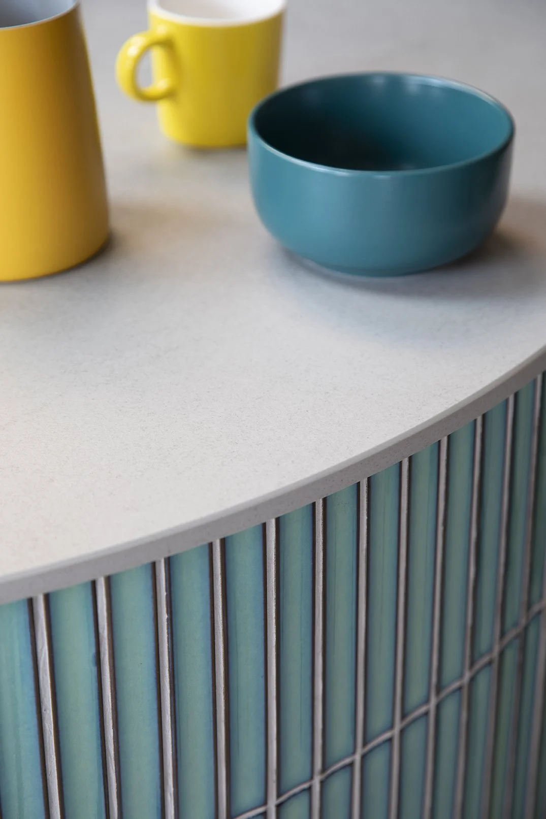 lose-up detail of the curved teal glazed finger tile cladding on the kitchen island in a North Shields family home by Carole Crowe Interior Design, showing the rounded grey stone worktop edge with yellow ceramic mugs and a teal ceramic bowl styled on