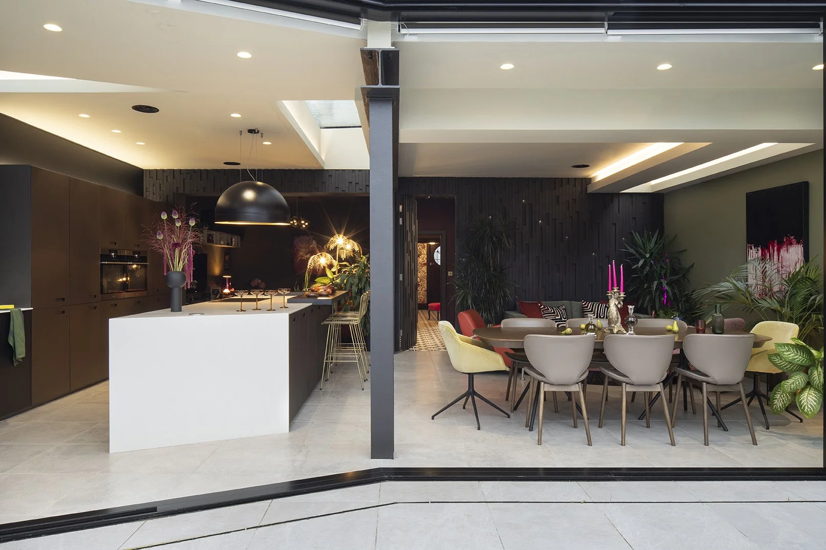 Hero view from garden through open bifold doors into open-plan kitchen and dining extension, showing white island, brown cabinets, black dome pendant, mixed velvet dining chairs and olive green feature wall, Queens Road Whitley Bay