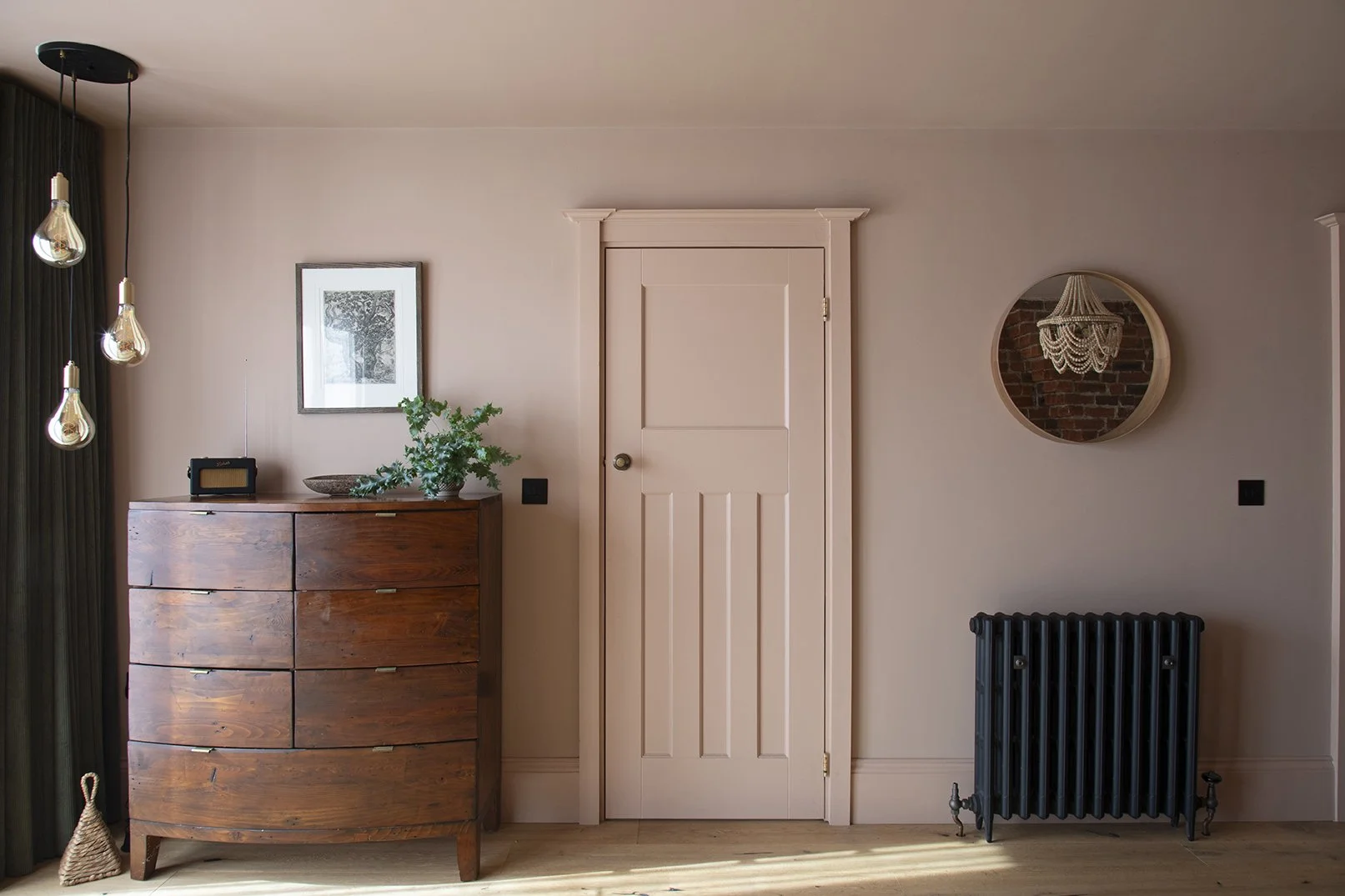 Loft bedroom in the Queens Road project by Carole Crowe Interior Design, Whitley Bay, showing a blush pink painted Victorian panelled door with a brass knob, a dark reclaimed wood bow-fronted chest of drawers with a Roberts radio and trailing ivy pla