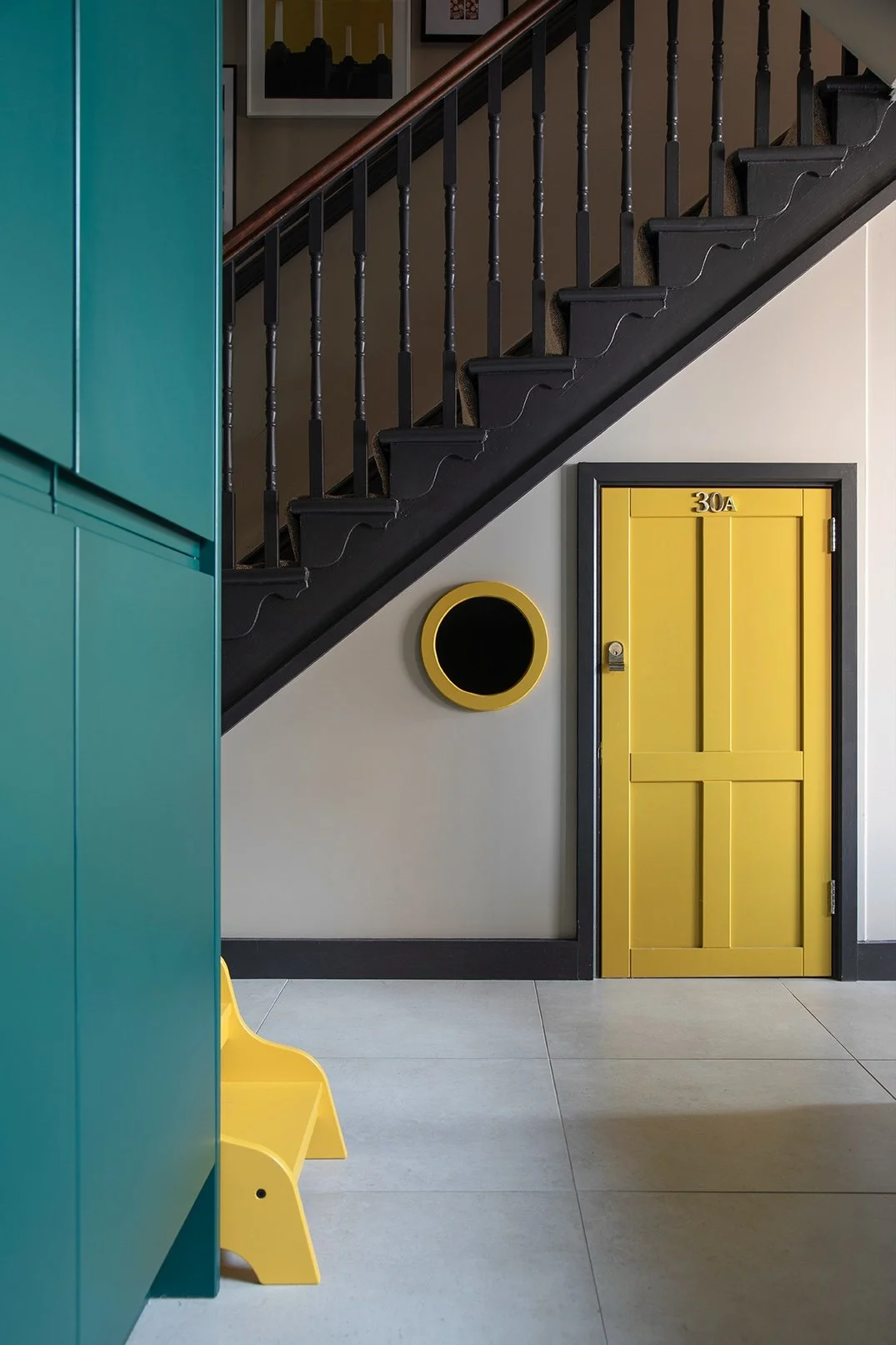 Close-up of the yellow painted under-stairs children's playroom door numbered 30A with a circular yellow-framed cat flap in a North Shields family home by Carole Crowe Interior Design, set against a painted black Victorian staircase with turned spind