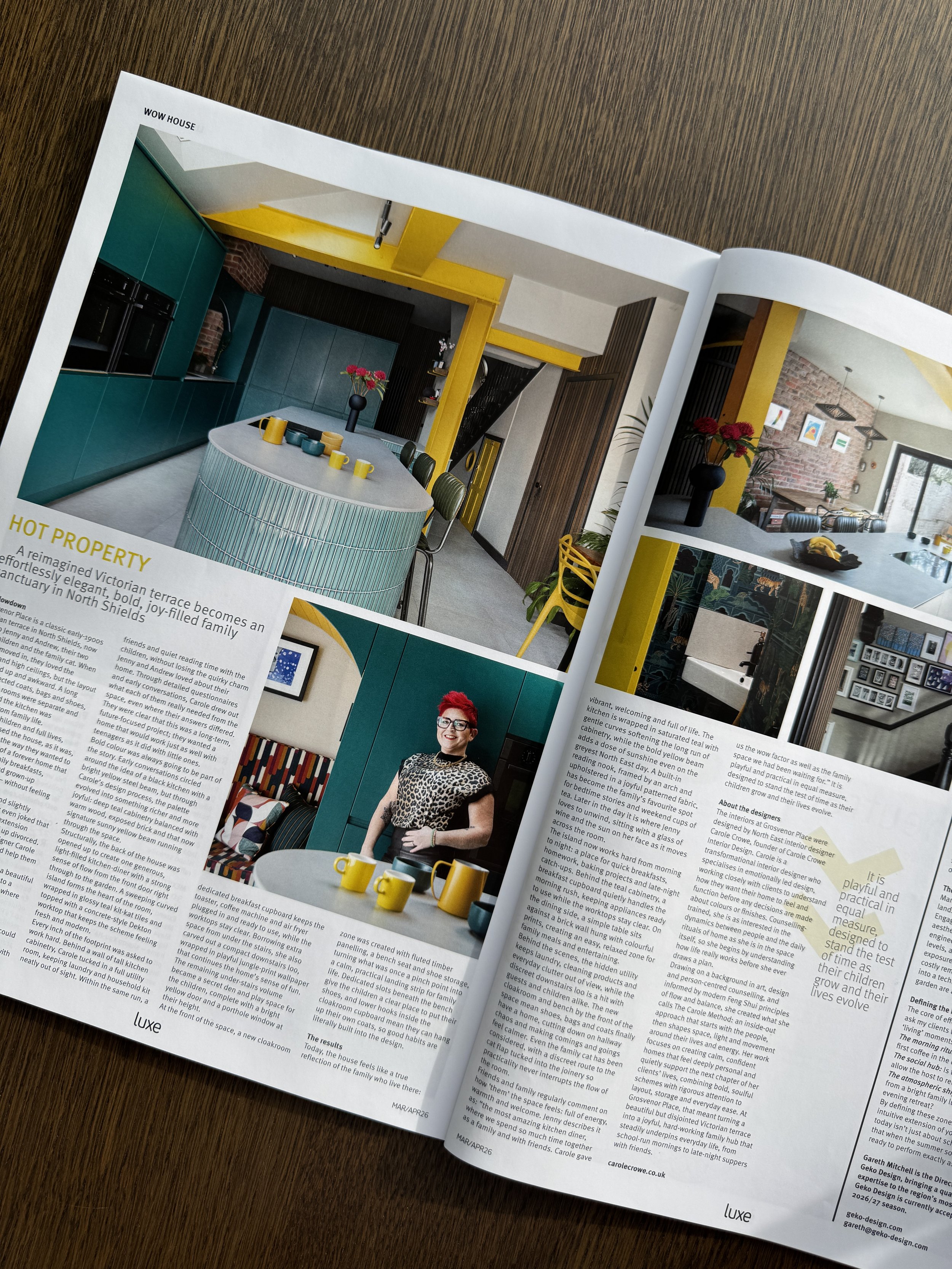 An open magazine showing pages with images of a modern kitchen and a woman with short red hair, glasses, wearing a leopard-print top, standing next to a round table with yellow cups and a tea set, on a wooden surface.