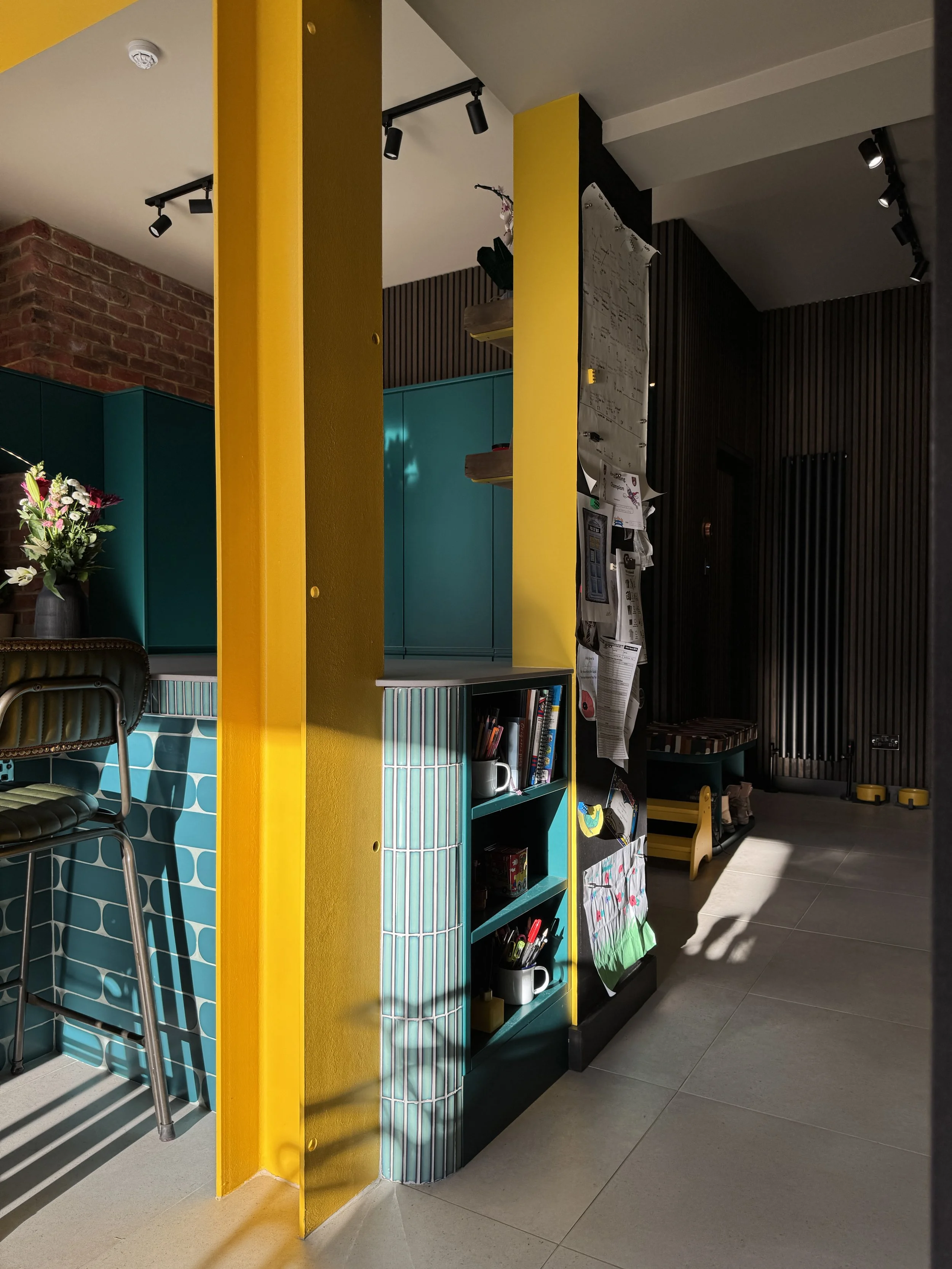 Candid everyday view of the open-plan kitchen in a North Shields family home designed by Carole Crowe Interior Design, showing the curved teal tiled island with open shelving, a yellow painted structural steel frame, a family notice board pinned with