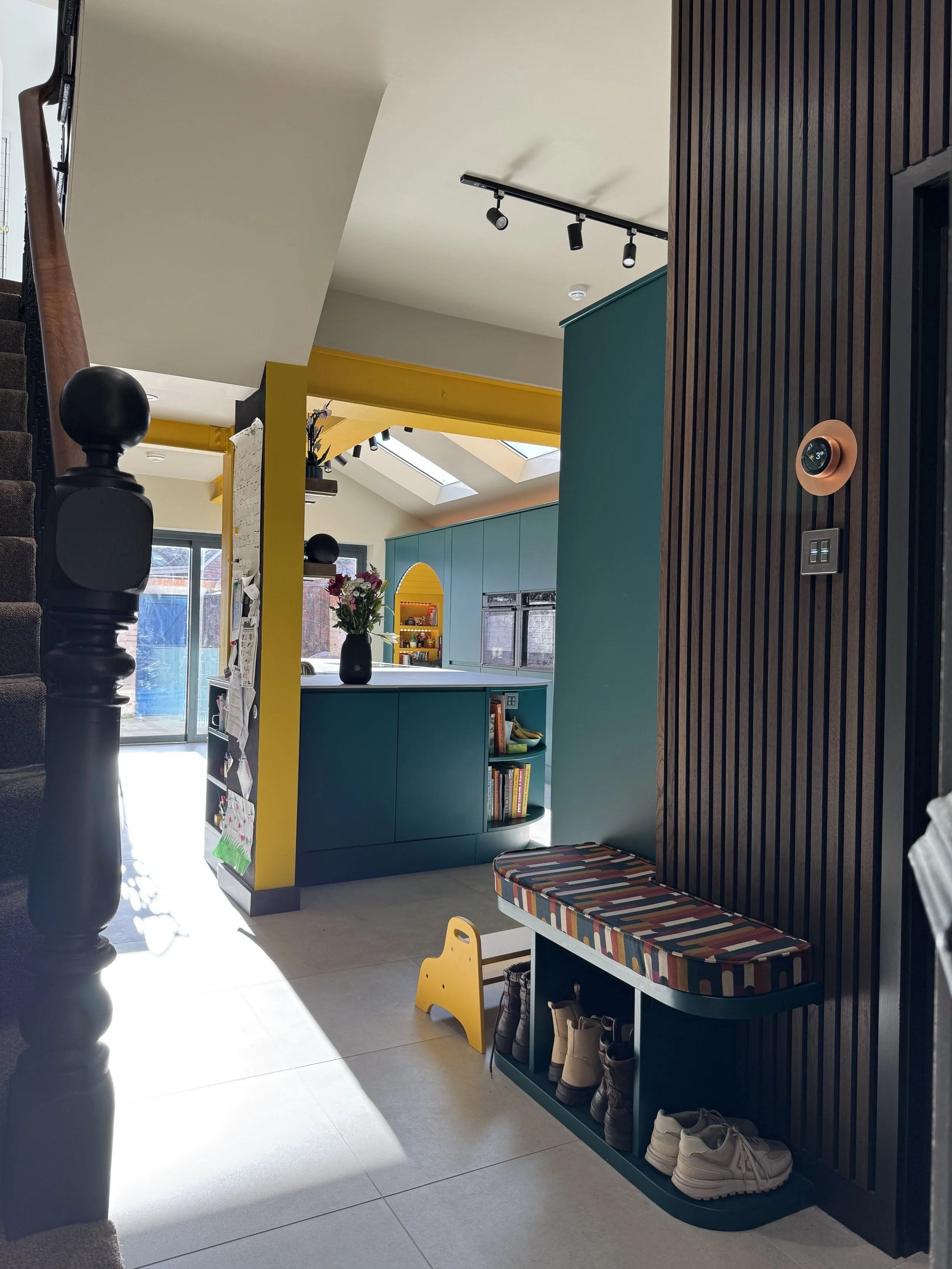View from the hallway and boot room through to the open-plan kitchen in a North Shields family home designed by Carole Crowe Interior Design, showing the teal shoe storage bench with colourful striped upholstered seat, a yellow children's step stool,