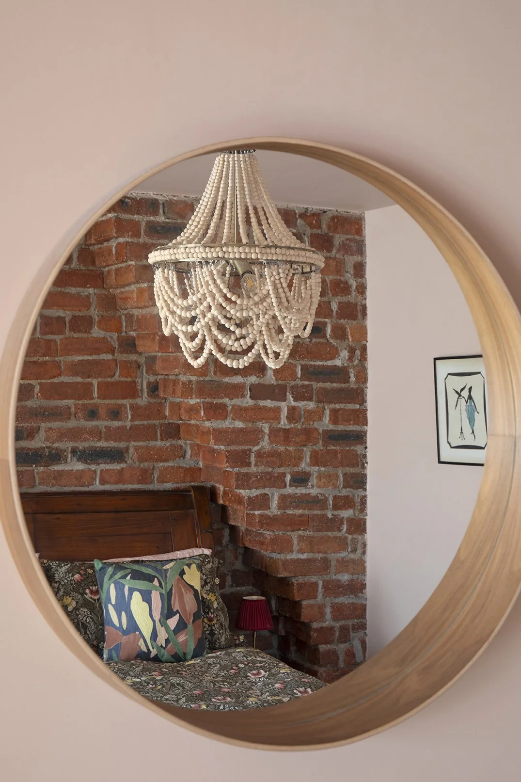 Round oak-framed mirror in the bedroom of the Queens Road project by Carole Crowe Interior Design, Whitley Bay, reflecting the cream wooden bead chandelier, the exposed red brick chimney breast wall, the dark wood sleigh bed with botanical print cush