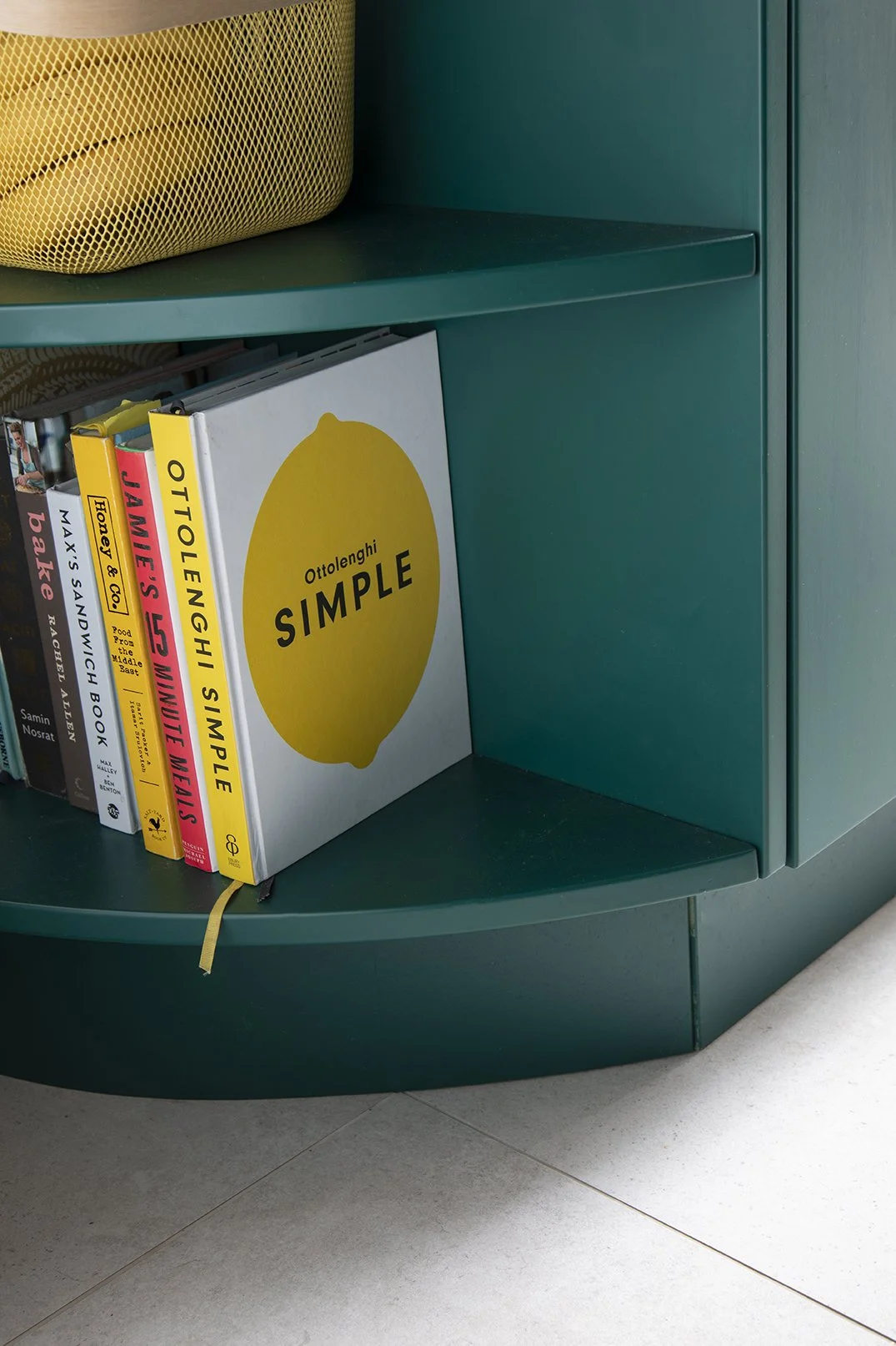 Detail of the curved open shelf at the base of the teal kitchen island in a North Shields family home designed by Carole Crowe Interior Design, styled with a collection of cookbooks including Ottolenghi Simple and Jamie's 5 Ingredient Meals, with a y