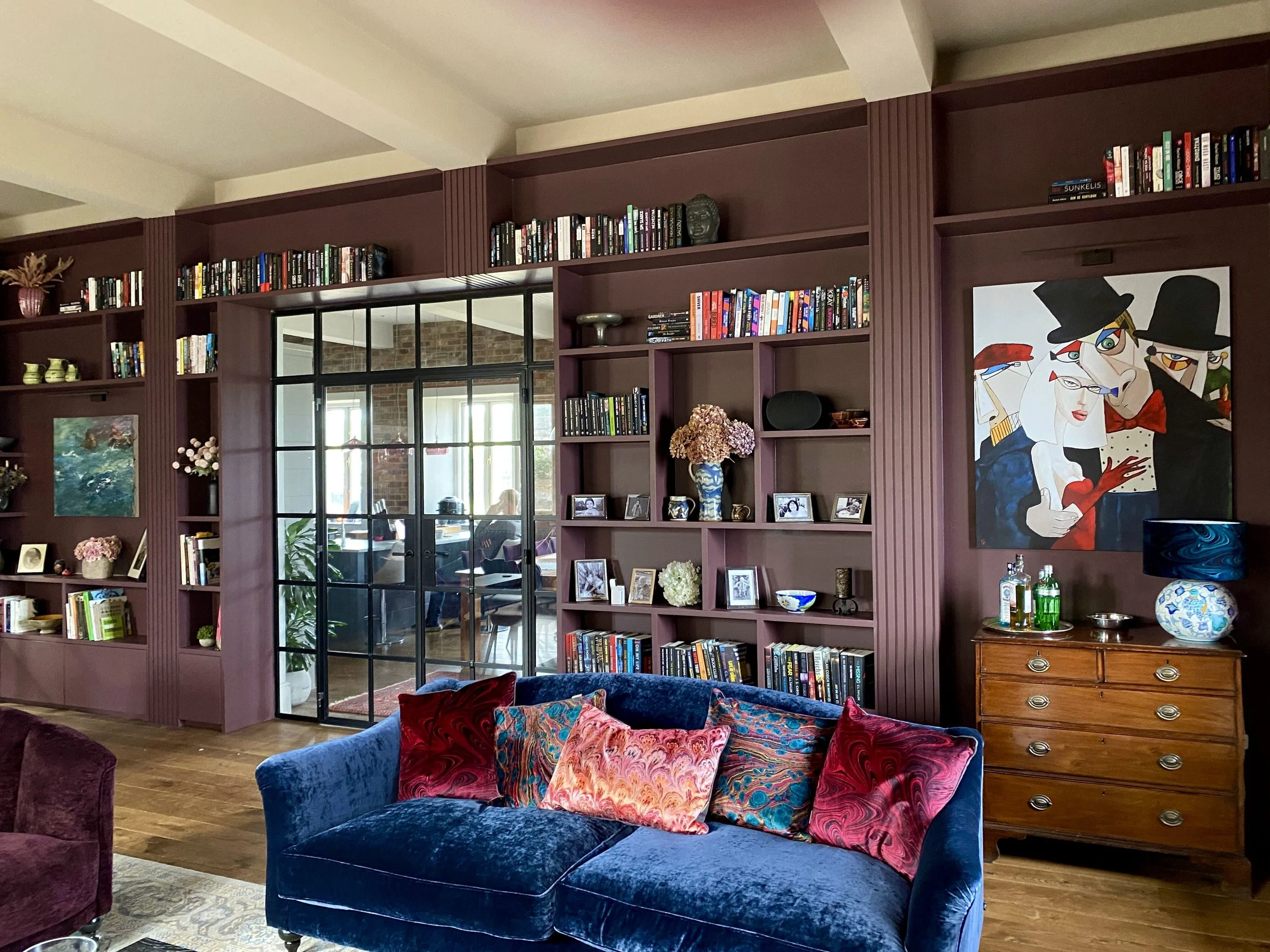 Living room with burgundy bespoke built-in library , colorful framed art, and velvet couch with feature pillows. luxury library living room for entertaining and relaxation, timeless contemporary classic design