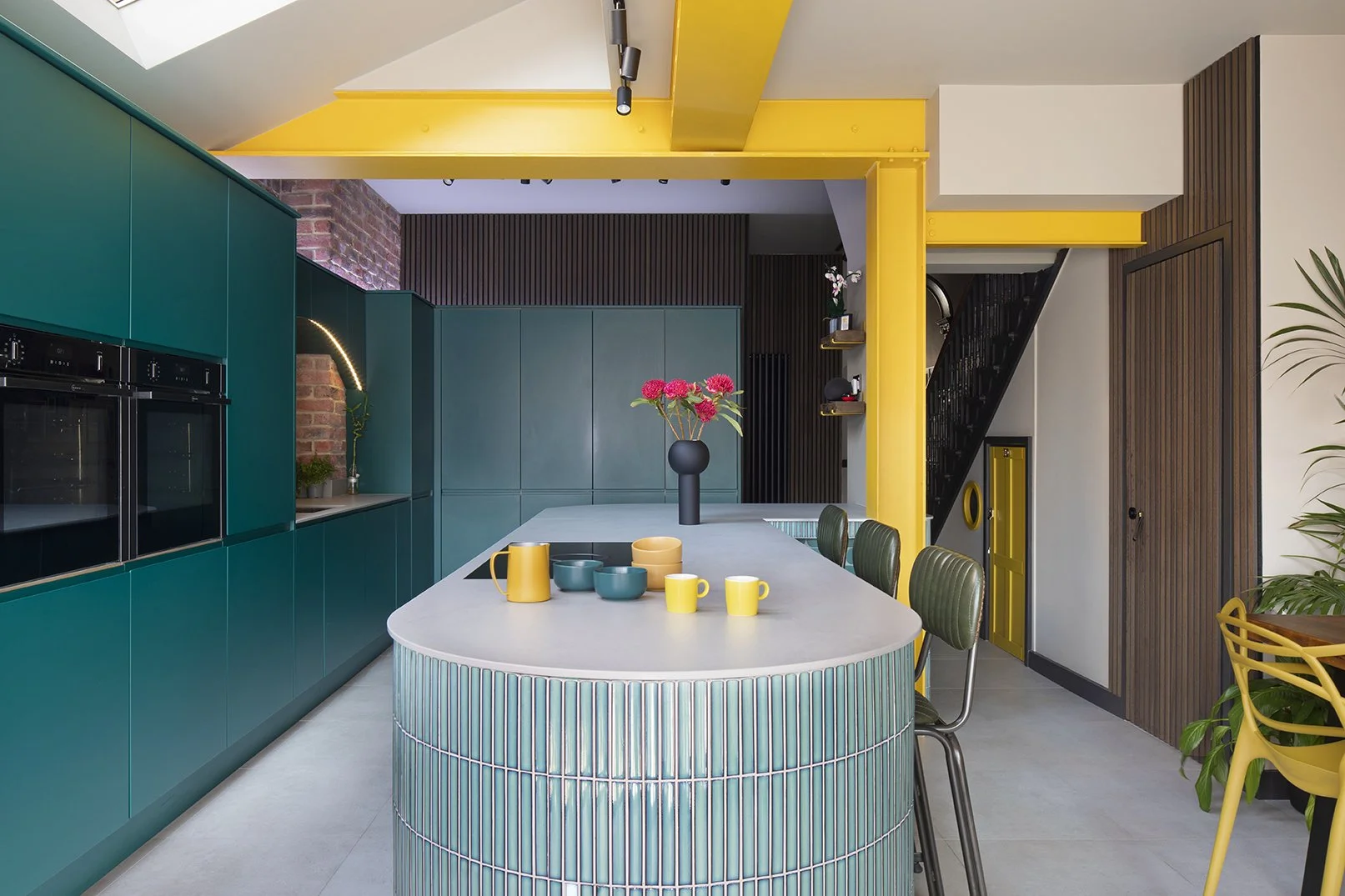 Detail of a bold yellow painted structural steel column in a North Shields kitchen designed by Carole Crowe Interior Design, set against a dark timber slatted door panel and exposed red brick wall, with a grey stone kitchen island worktop in the fore