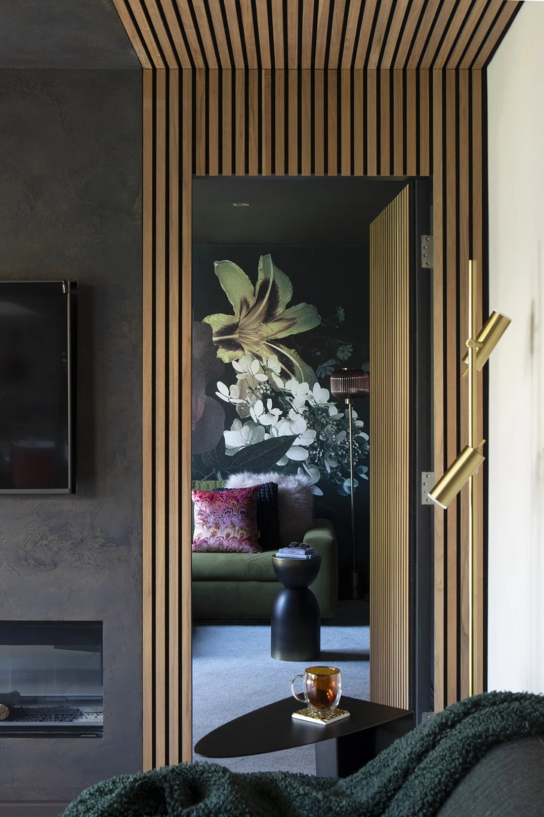 Close view through oak wood slat framed concealed snug doorway revealing dark botanical mural, olive sofa and brass adjustable wall light, Lytham Road Tynemouth