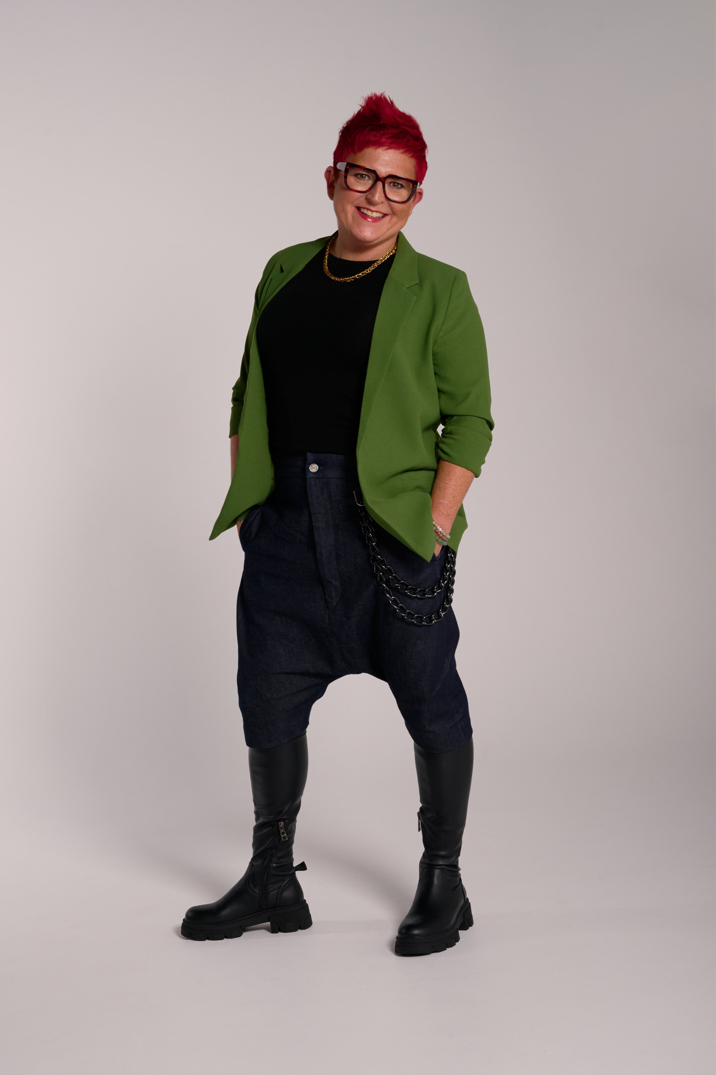 A woman with short, spiky red hair, wearing glasses, a green blazer, black top, dark denim shorts, chunky black thigh-high boots, and gold jewelry, standing casually against a plain background. Carole Crowe Transformational Interior Designer.