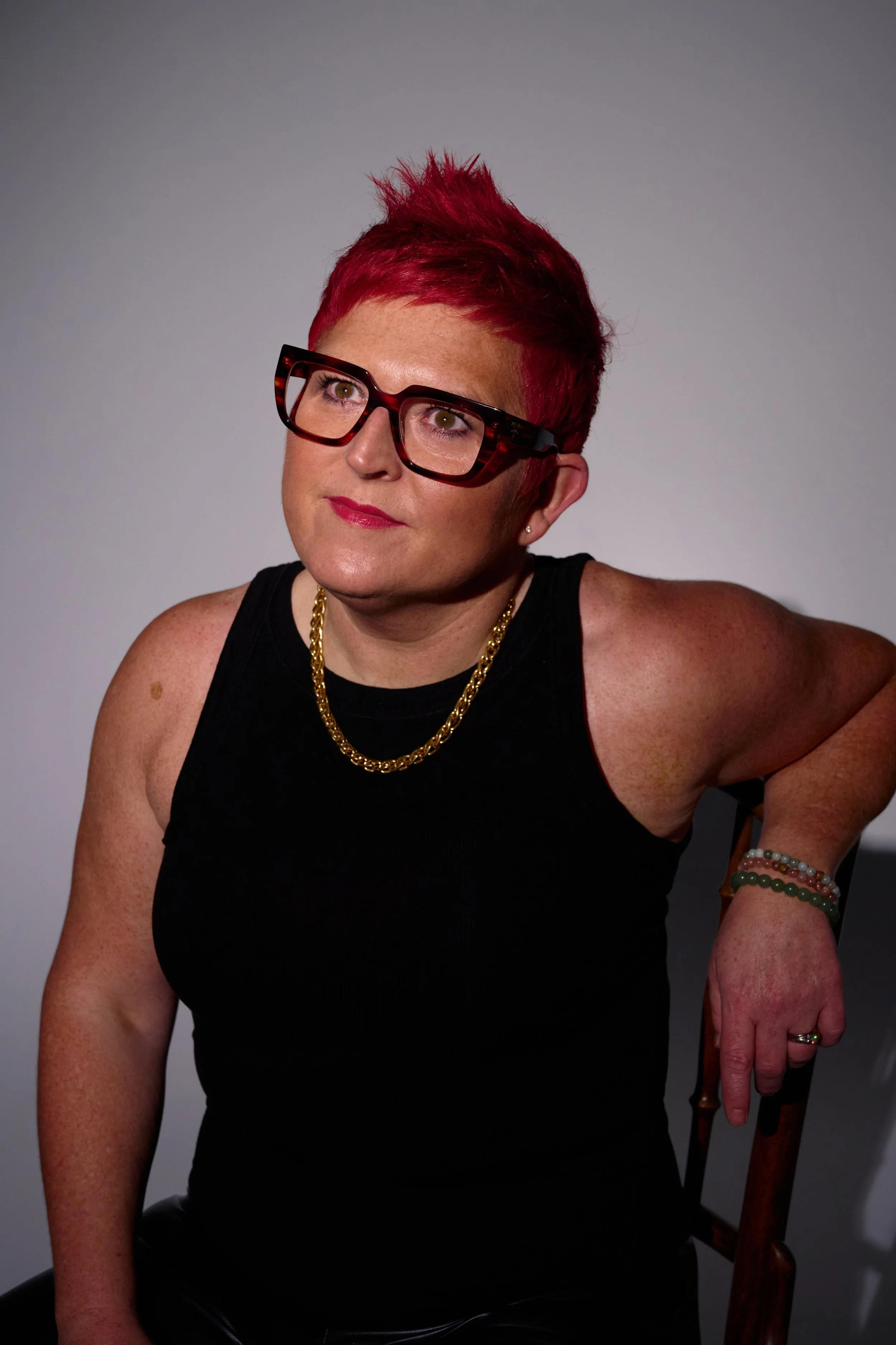 A woman with short red hair, wearing large black glasses, a black tank top, and gold jewelry, sitting on a wooden chair against a plain gray background.  Carole Crowe Transformational Interior Designer.