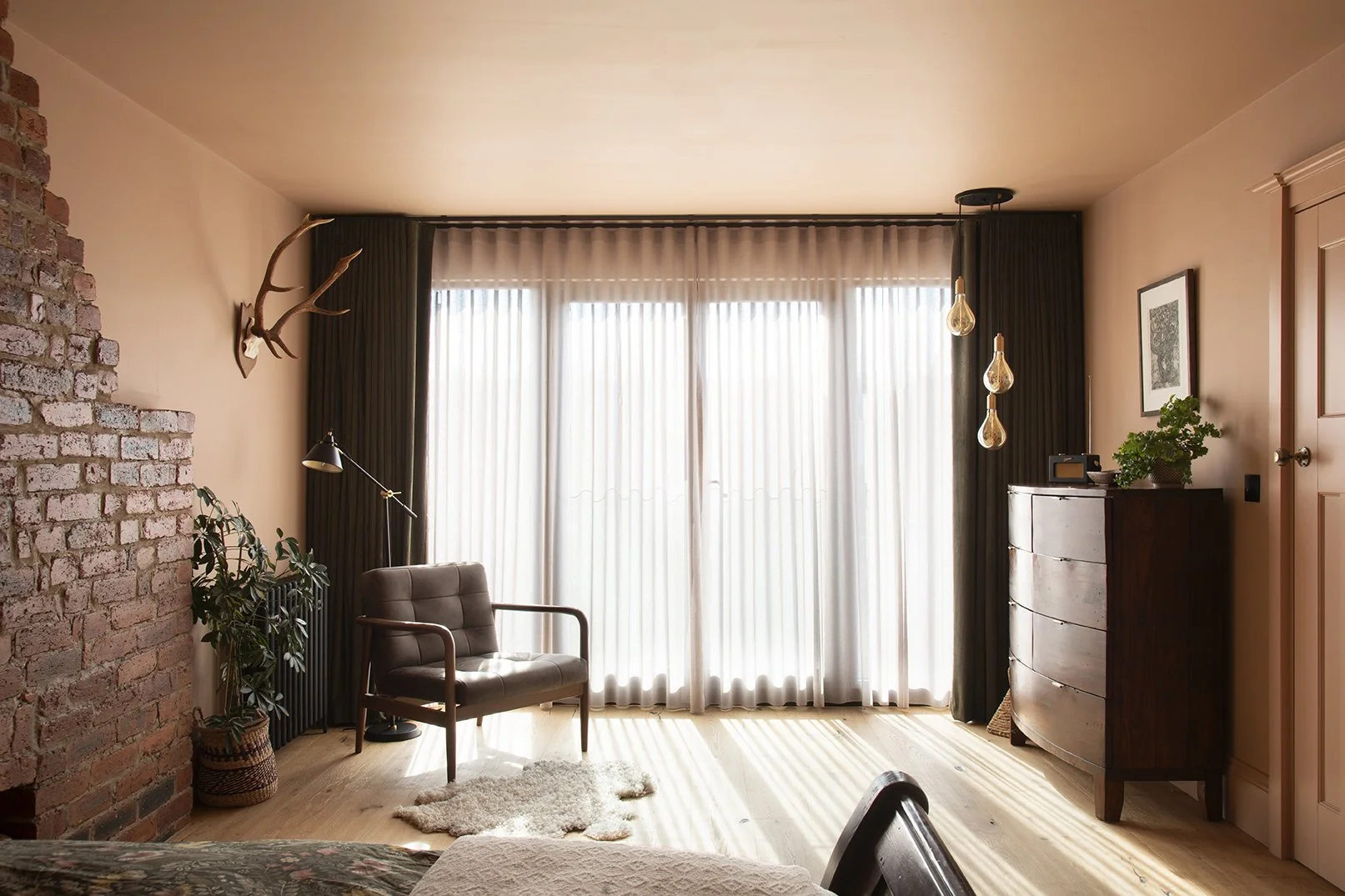 Full overview of the loft bedroom in the Queens Road project by Carole Crowe Interior Design, Whitley Bay, showing floor-to-ceiling sheer white and olive corduroy curtains across a wide black-framed picture window, a mid-century dark grey tufted armc