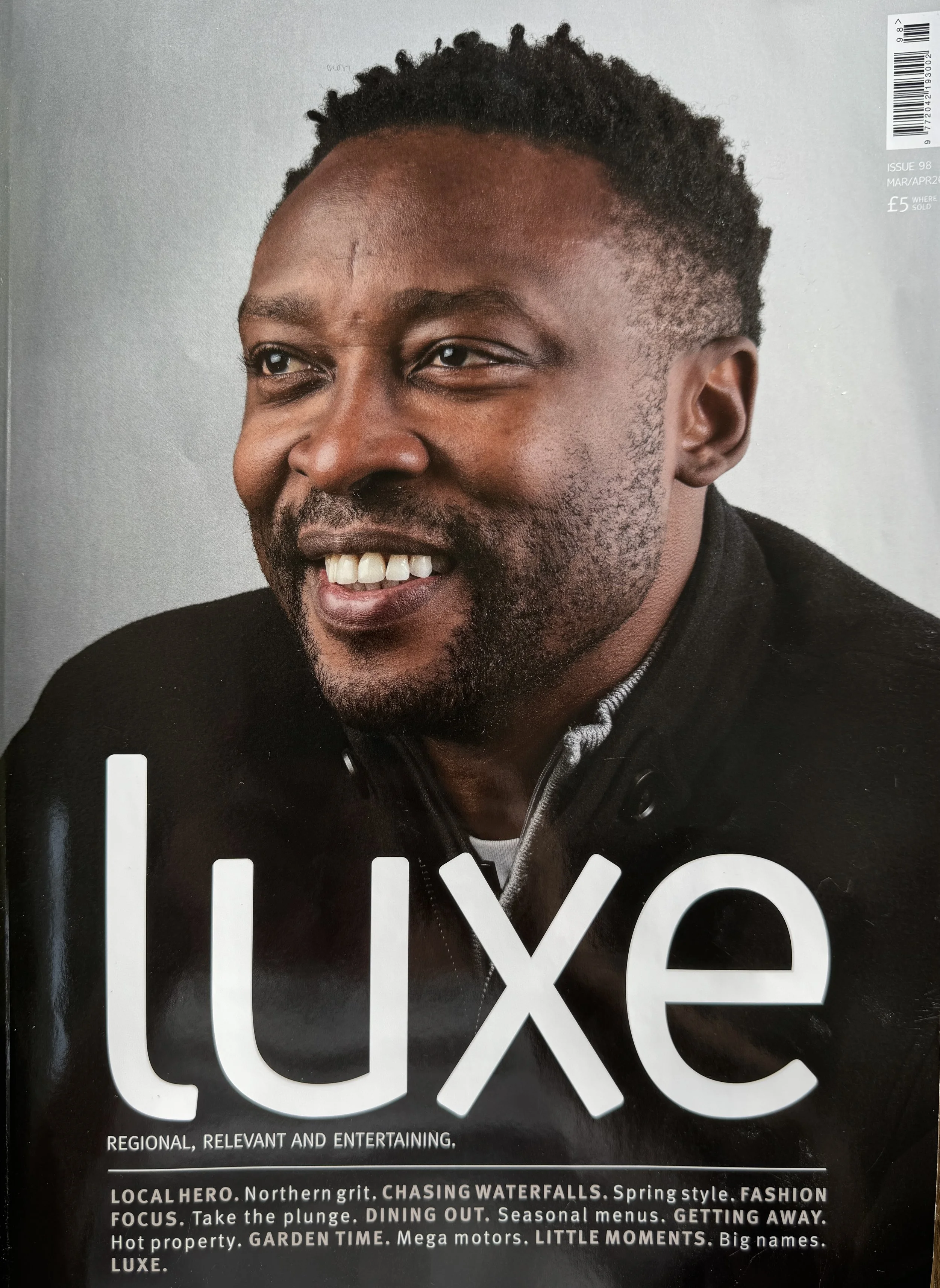 Close-up of a magazine cover featuring a smiling man with short, curly hair and a beard, wearing a black jacket, with the word "LUXE" in large white letters.
