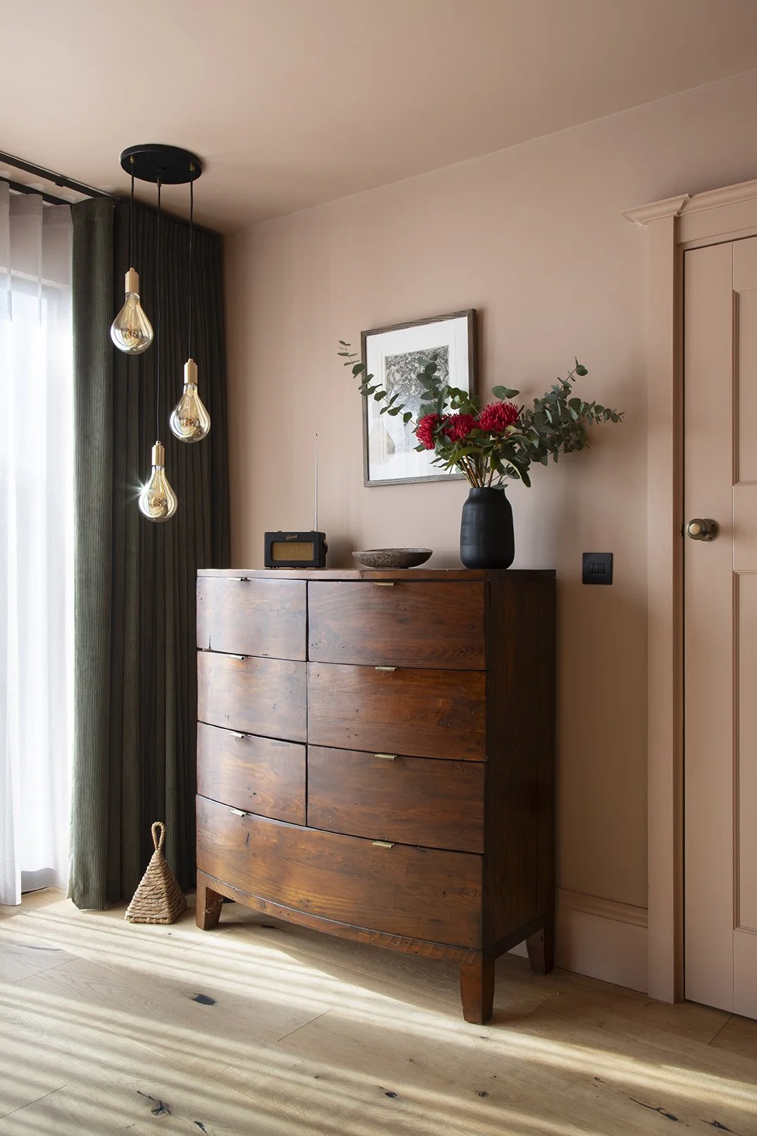 Loft bedroom in the Queens Road project by Carole Crowe Interior Design, Whitley Bay, featuring a dark reclaimed wood bow-fronted chest of drawers styled with a matte black vase of red waratah flowers and eucalyptus, a Roberts radio, a stoneware bowl