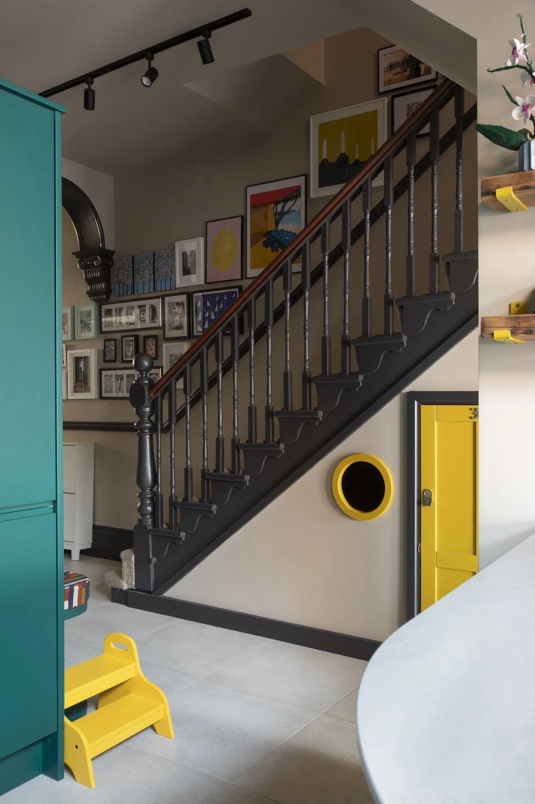Family kitchen designed by Carole Crowe Interior Design in North Shields, showing a run of floor-to-ceiling teal handleless cabinets with integrated double ovens, a curved tiled kitchen island with olive green leather studded bar stools, a yellow pai