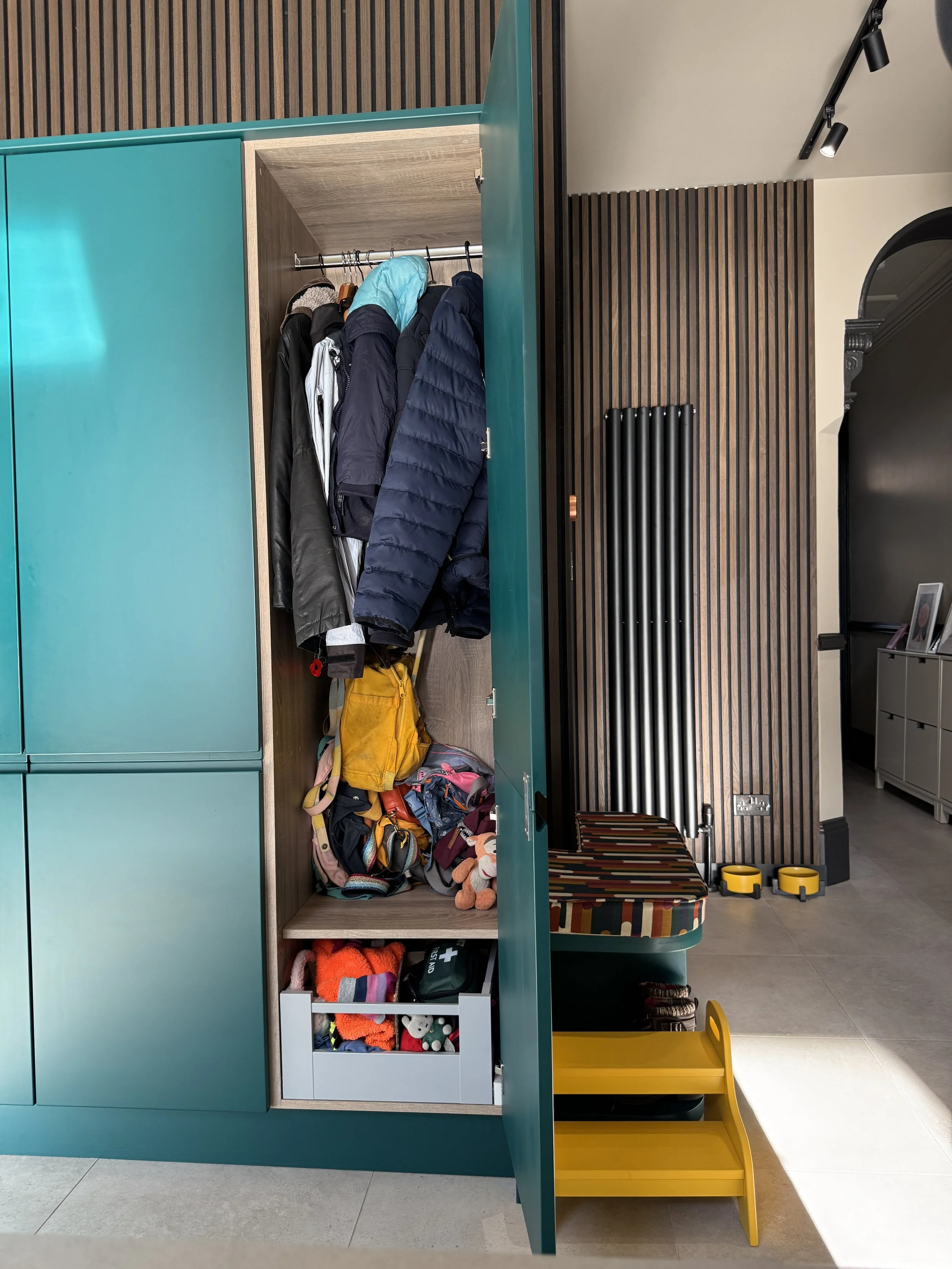 Open teal painted coat cupboard in the hallway of a North Shields family home designed by Carole Crowe Interior Design, showing a fully fitted interior with hanging rail for coats and jackets and a pull-out drawer below, alongside the colourful strip