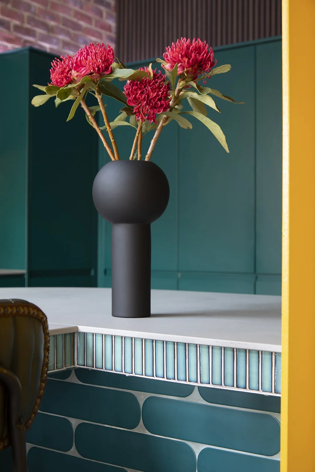 Styling detail on the kitchen island of a North Shields family home designed by Carole Crowe Interior Design, featuring a sculptural matte black mushroom-shaped vase with bold pink waratah flowers, set on a grey stone worktop above a curved teal glaz