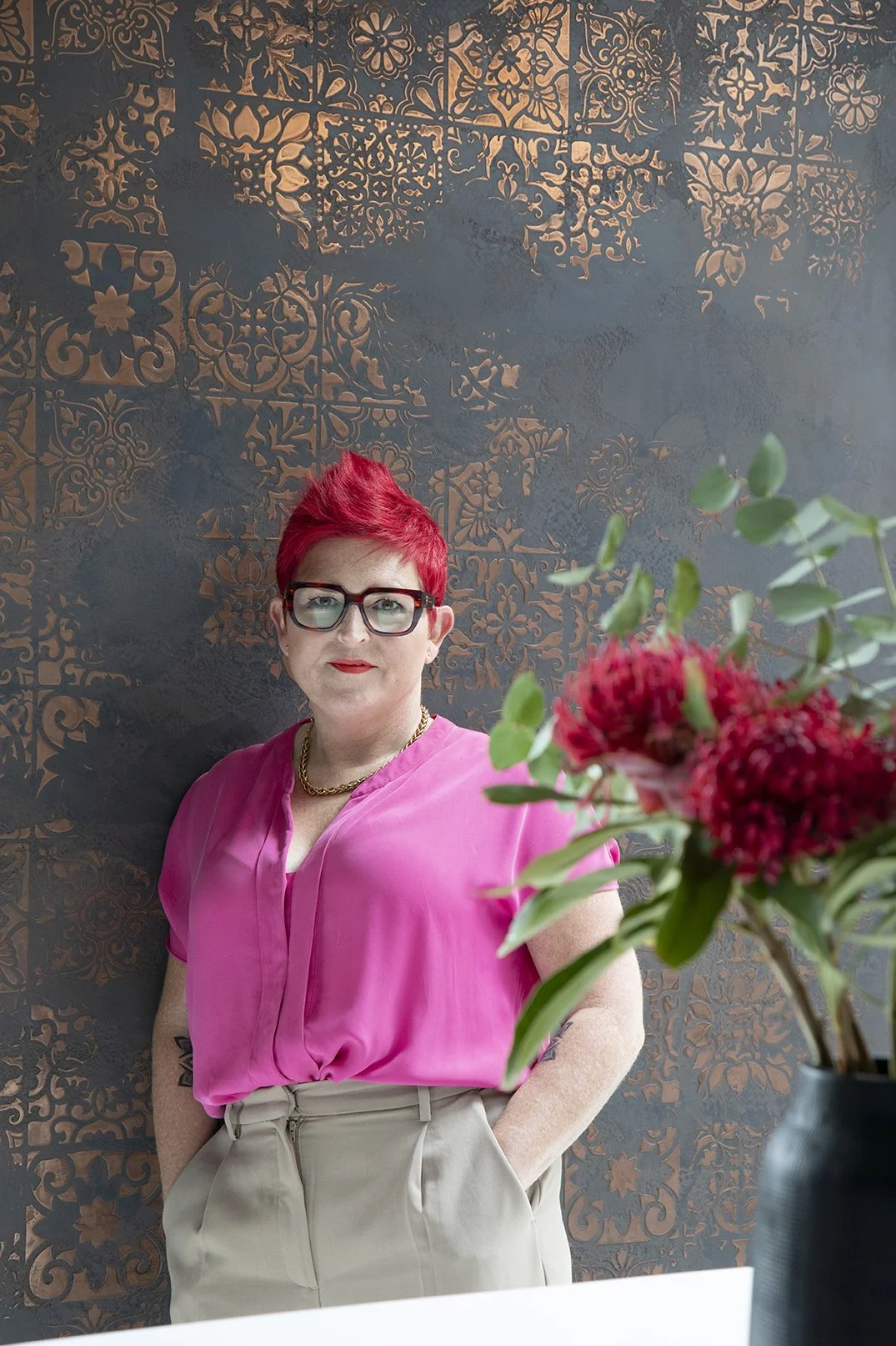 A woman with short red hair, glasses, and red lipstick stands against a decorative patterned wall. She is wearing a pink blouse and beige pants, with a gold chain necklace. A vase with red flowers and green leaves is in the foreground. Carole Crowe