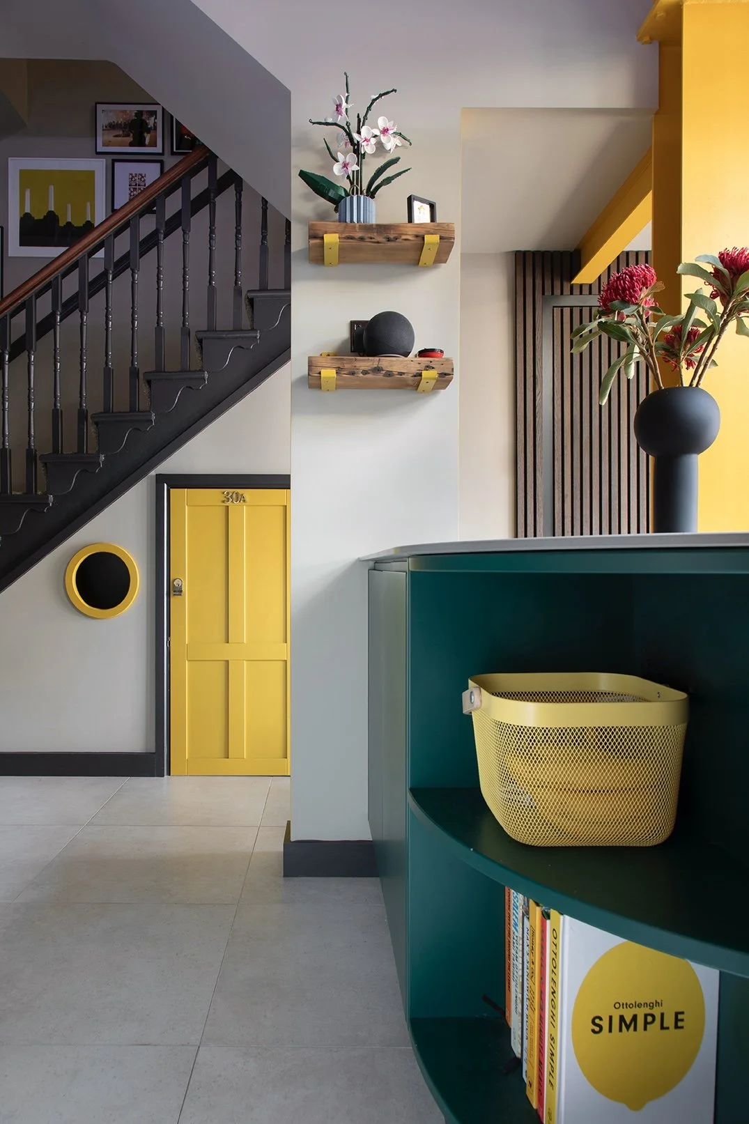 View from the kitchen island towards the staircase hall in a North Shields family home designed by Carole Crowe Interior Design, showing the curved teal island with open cookbook shelving and yellow mesh storage basket, a painted black Victorian stai