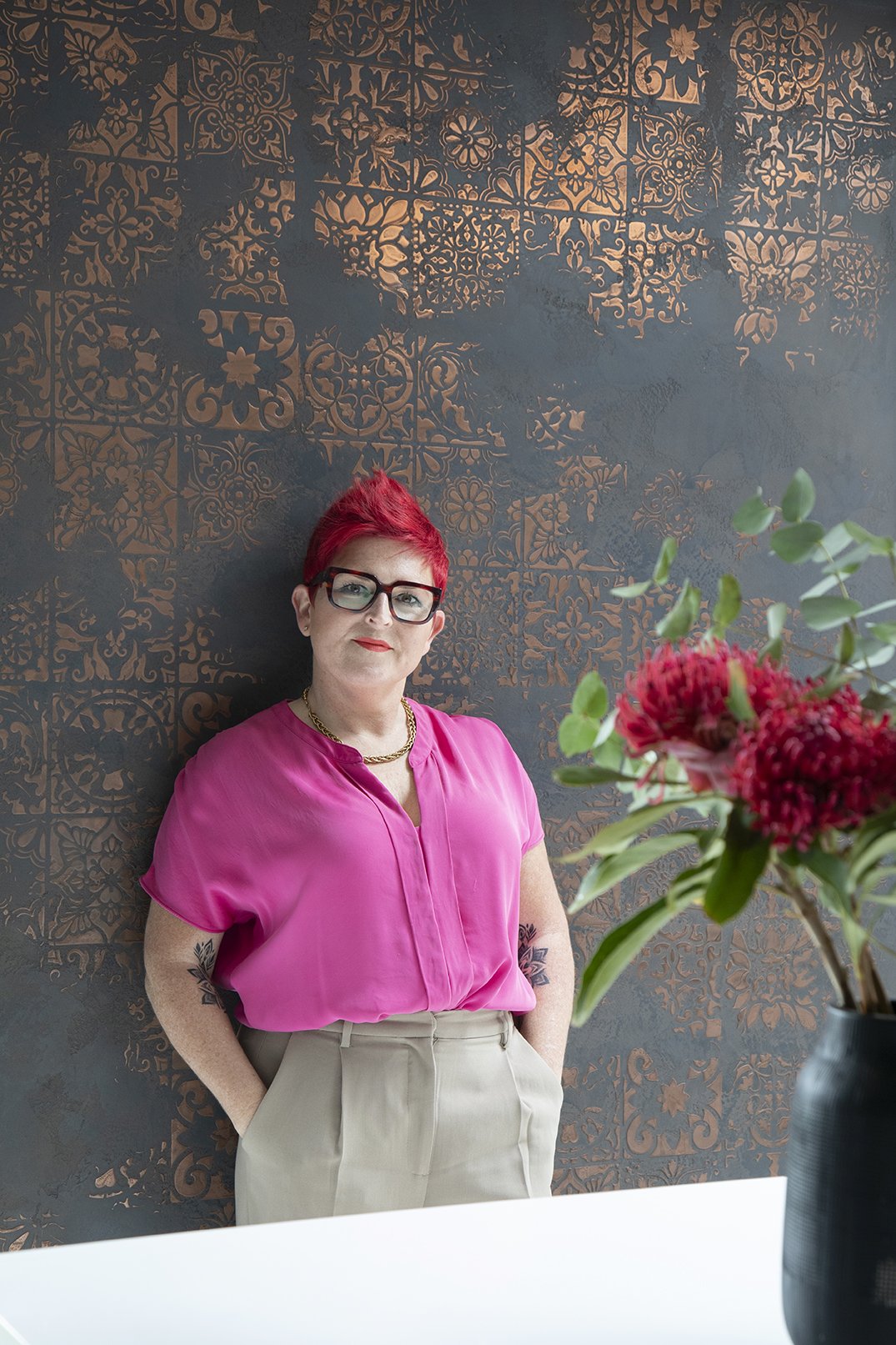 A woman with bright red hair, glasses, and tattoos wearing a pink blouse and beige pants standing against a dark wall with floral patterns, with a vase of red flowers in the foreground.