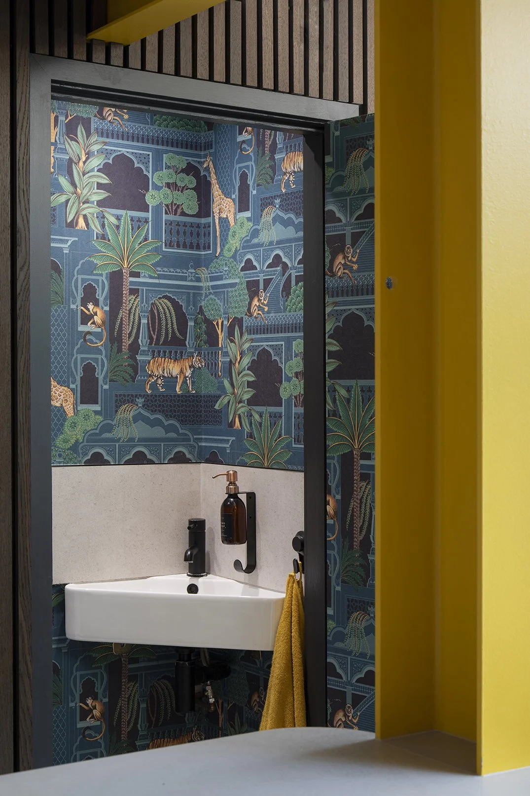 Downstairs cloakroom viewed through the open yellow painted door frame in a North Shields family home by Carole Crowe Interior Design, with dramatic floor-to-ceiling jungle print wallpaper featuring tigers, giraffes, monkeys and tropical palms within