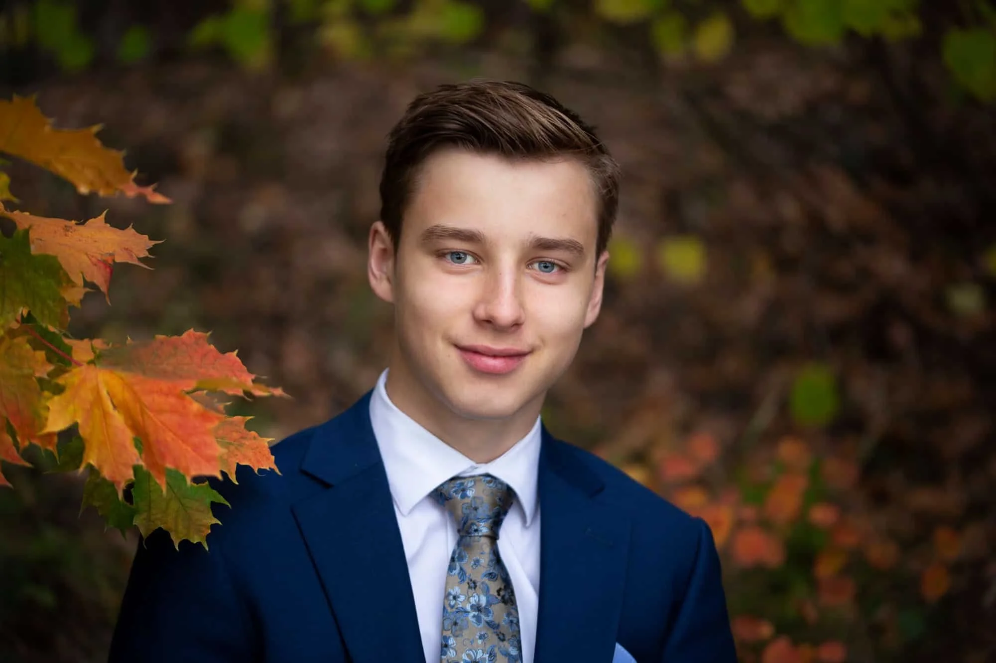 Konfirmant i blå dress ser i kameraet og på den ene siden stikker ei grein frem med blader i sterke høstfarger